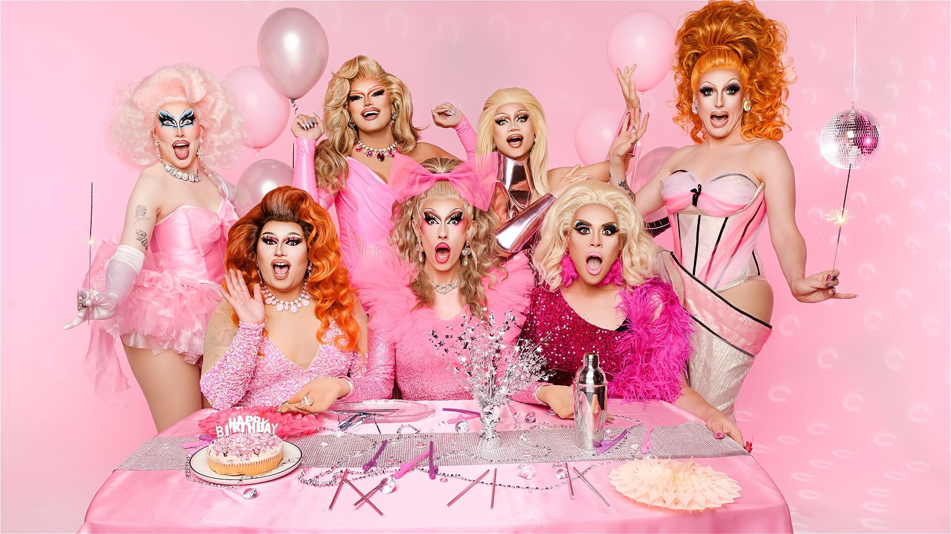 Seven drag queens in glamorous, colorful outfits pose at a festive pink-themed table with balloons, a cake, and party decorations. The background is pink, creating a vibrant, celebratory atmosphere.