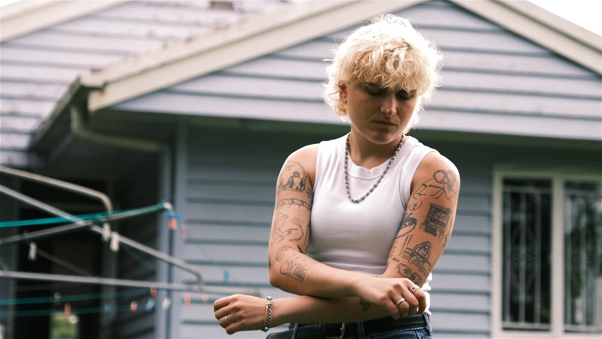 A person with short blonde hair and tattooed arms stands outside a light blue house, looking down while adjusting their sleeve. They wear a white sleeveless top, necklace, and jeans.