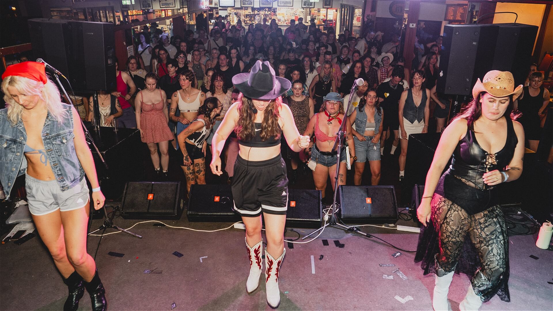 Three women in cowboy hats and Western outfits dance on stage, while a large crowd of people watches from the floor of a lively, indoor venue decorated with posters and memorabilia.