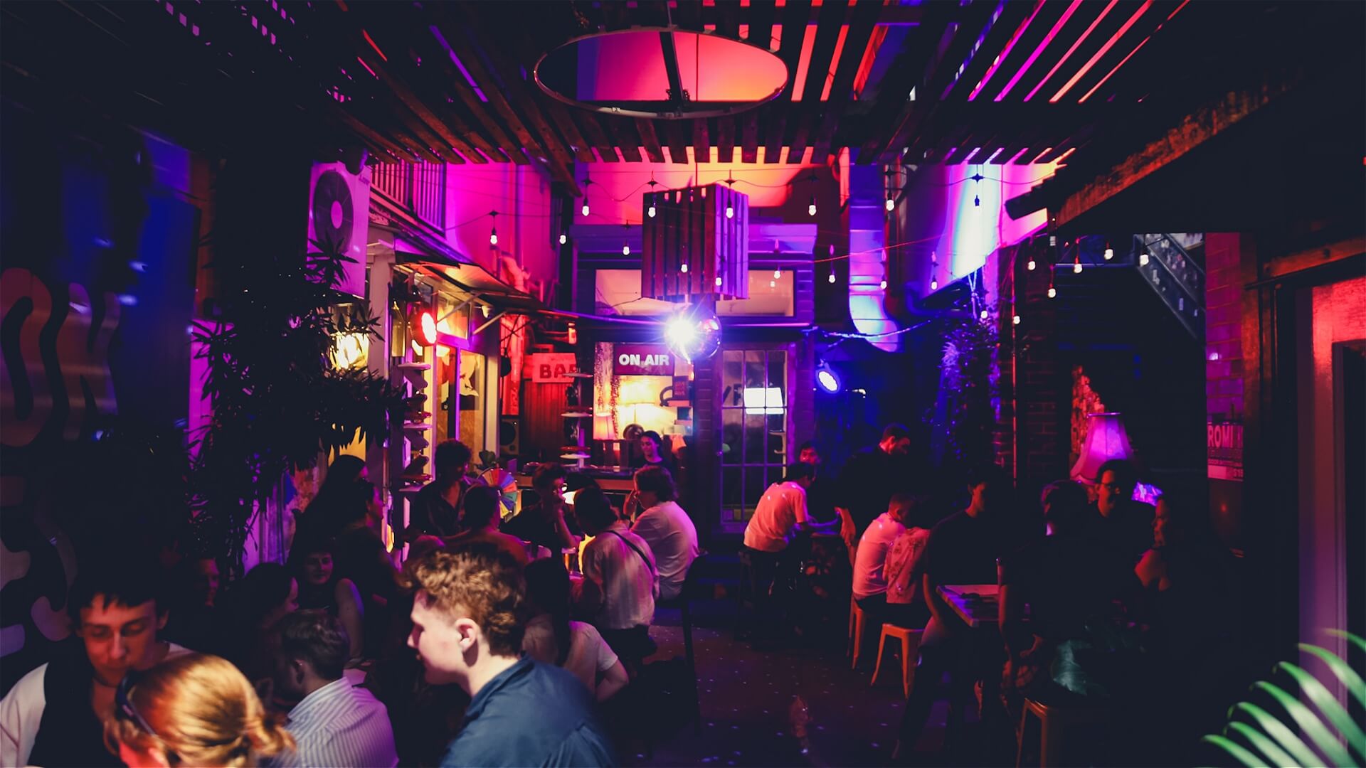 A vibrant, crowded bar with colorful neon lights and people sitting at tables, talking and socializing in a lively, indoor atmosphere.