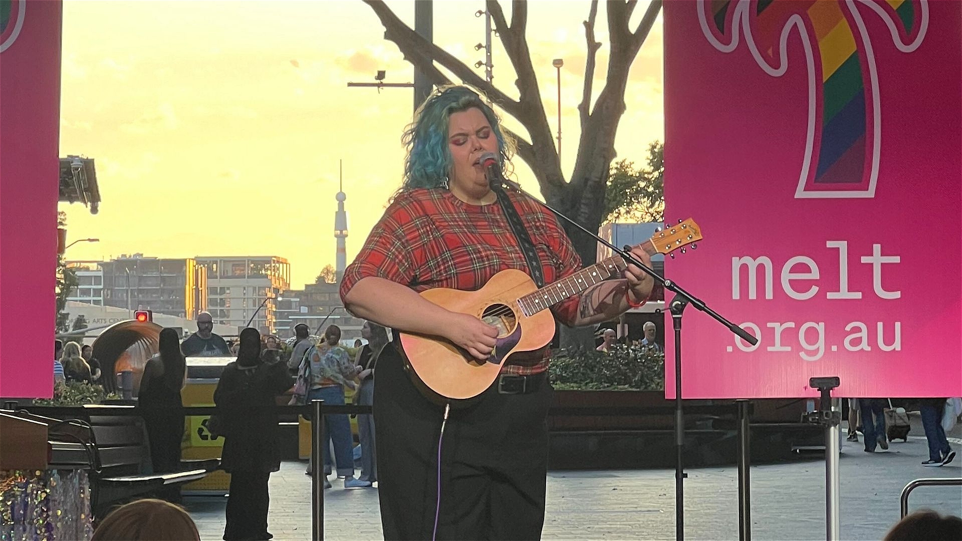 A person with blue hair plays an acoustic guitar and sings into a microphone on an outdoor stage. Behind them are large pink banners with rainbow palm tree logos and the text melt.org.au. People and buildings are visible in the background.