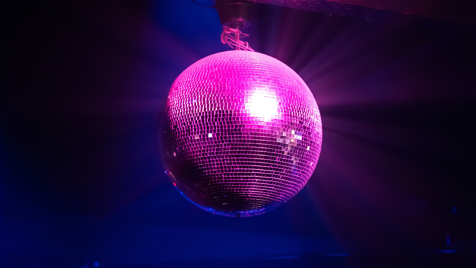 A shiny disco ball reflecting pink and purple light beams hangs from the ceiling in a dark room, creating a vibrant and festive atmosphere.