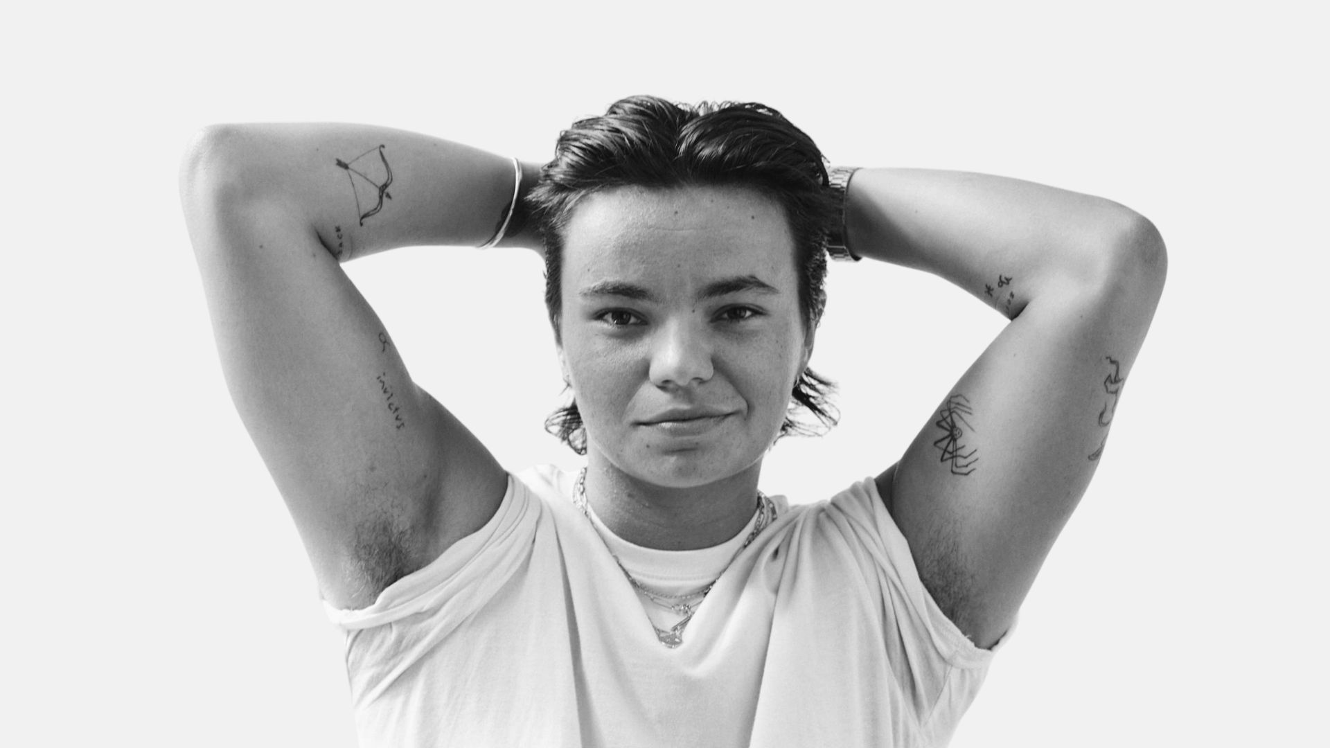 Black and white photo of Zoe Terakes sitting with hands behind their head, wearing a white t-shirt and jeans. With short dark hair and visible tattoos, they look directly at the camera.