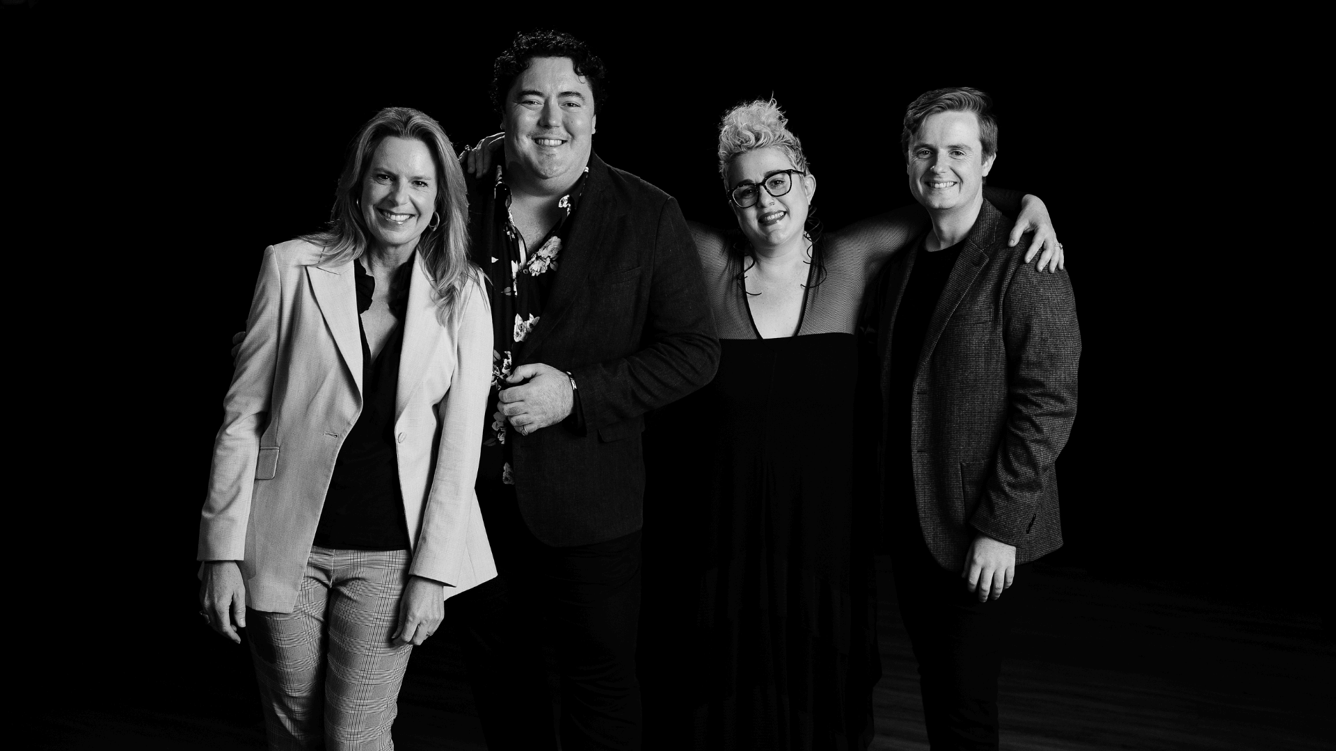 Four people stand together smiling against a black background. Two women are on the left, and two men are on the right. They are dressed in business-casual attire and appear happy and friendly.