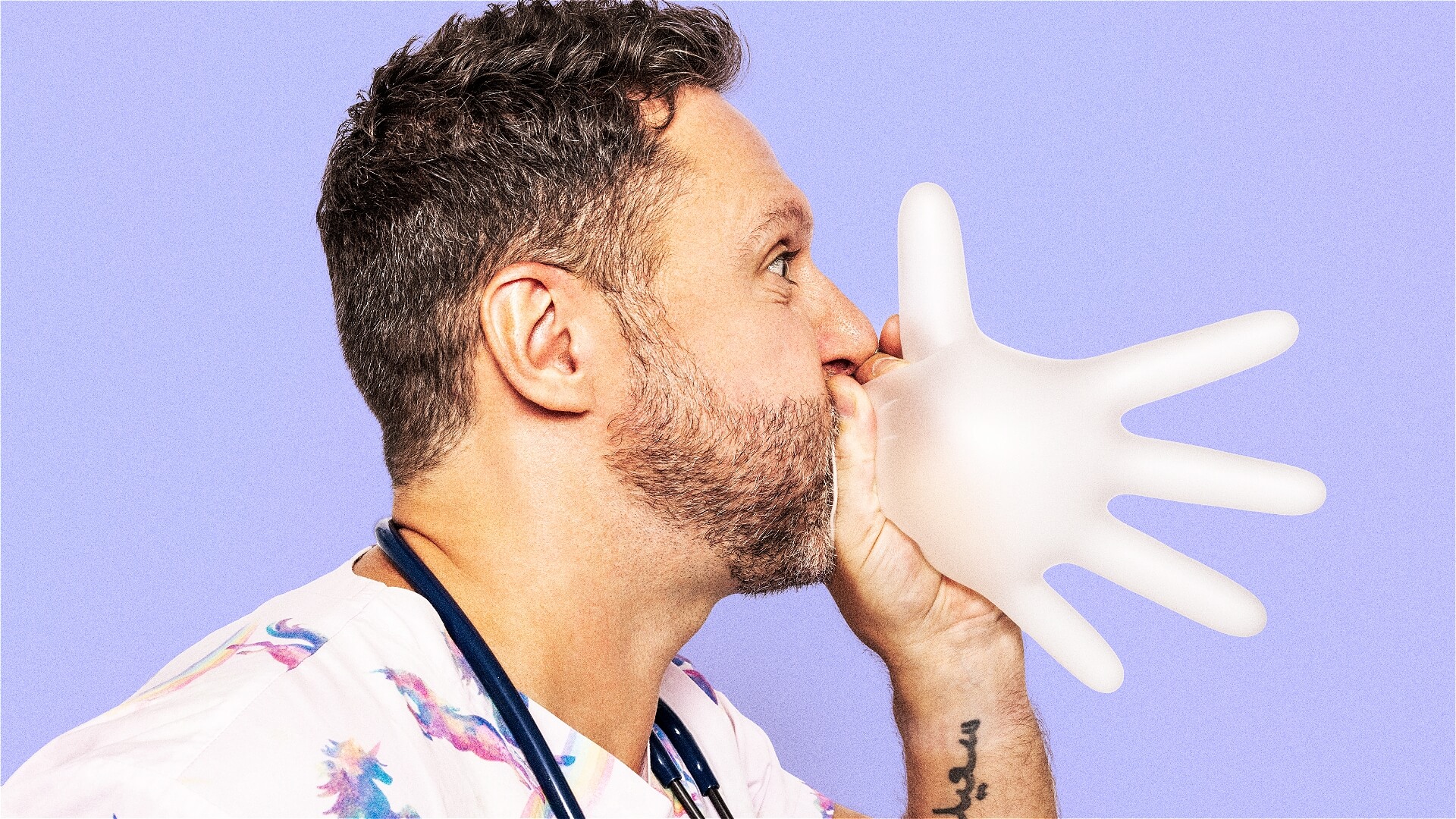 A man with a beard blows air into a white latex glove, inflating it like a balloon. Wearing fun scrubs and a stethoscope, he stands against a purple background.