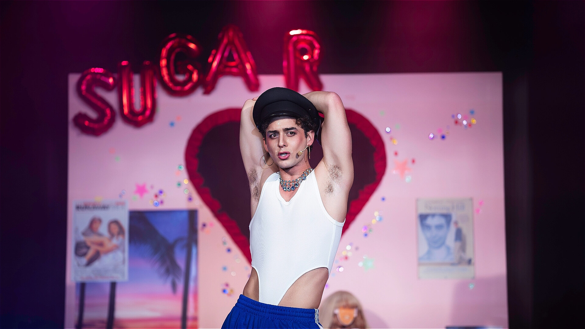 A performer in a white cut-out tank top and blue pants poses on stage with arms raised. Behind them is a heart-shaped backdrop, pink lighting, and balloons spelling SUGAR. Posters and sparkles decorate the background.