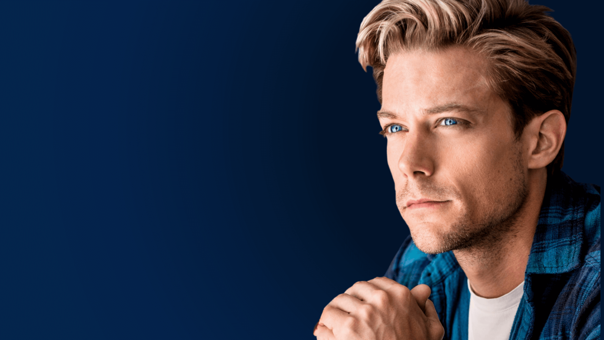 A young man with blond hair and blue eyes gazes thoughtfully into the distance, resting his chin on his clasped hands. He wears a blue shirt, and the background is a solid dark blue.