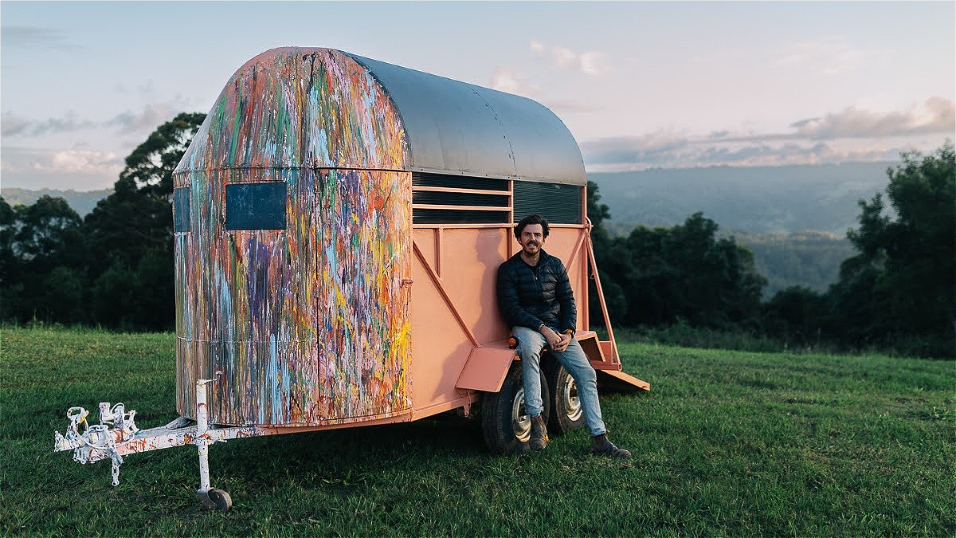A man sits on the edge of a painted trailer in a grassy field with trees and mountains in the background, relaxing in an outdoor setting under a cloudy sky.