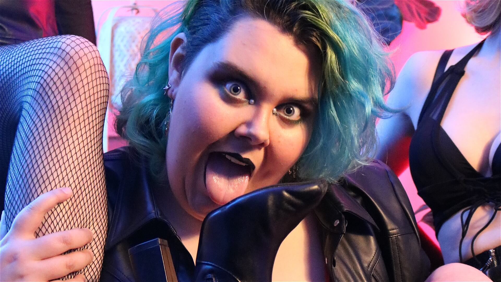 A person with teal hair and dramatic makeup sticks out their tongue toward a high-heeled boot, surrounded by people in fishnet stockings and black outfits, in a colorful, playful setting.
