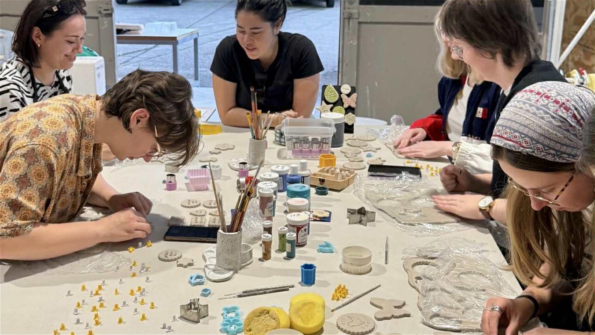 Five women sit around a table covered with clay, paint, brushes, and crafting tools, focusing on creating and decorating small clay objects in a well-lit studio space.
