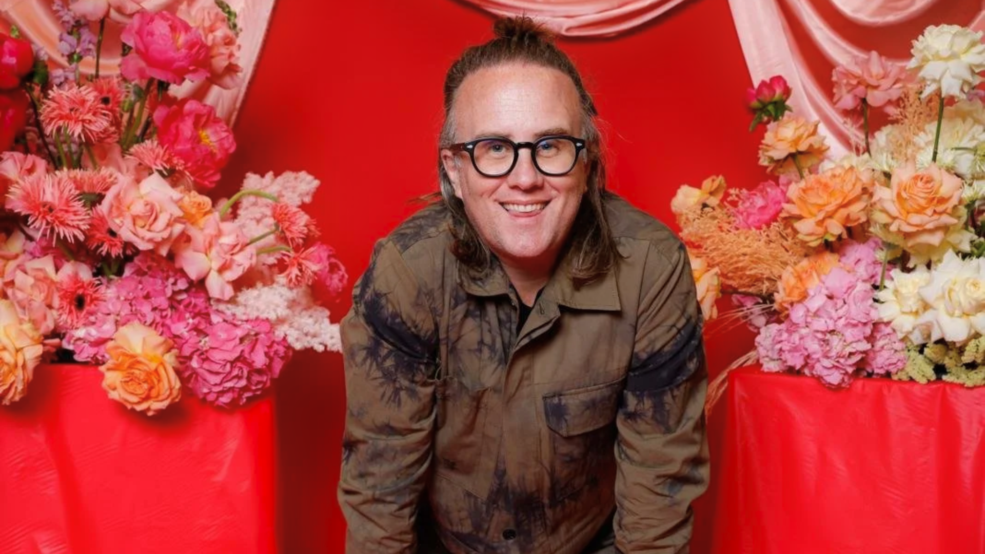 A person with long hair and glasses smiles, leaning forward in front of a bright red background decorated with pink and orange flowers arranged on both sides.