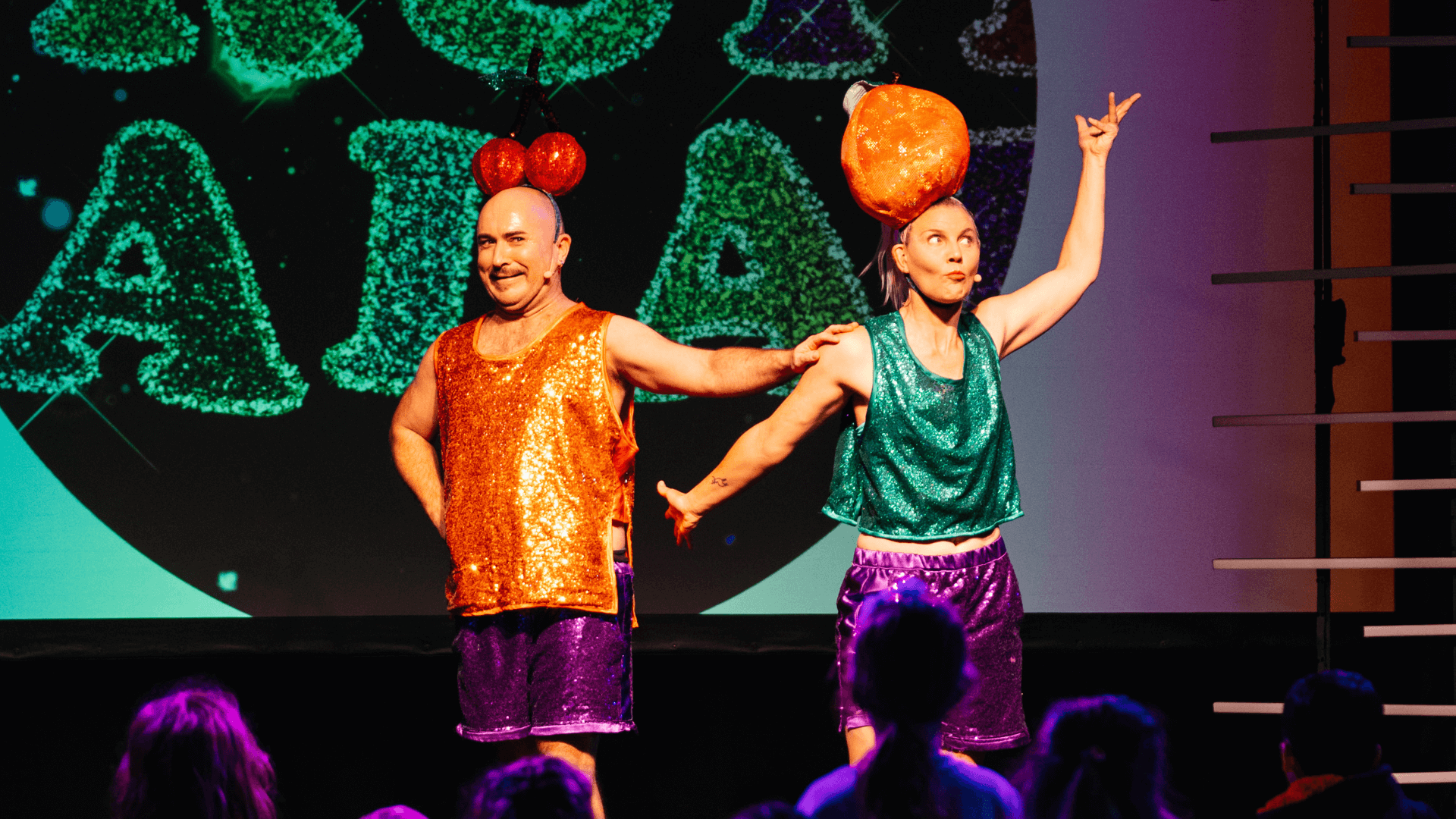 Two performers in sparkly outfits stand on stage. One wears an orange top with cherry headpiece, the other a green top with a large orange fruit headpiece, engaging with an audience of children. Bright, playful background.