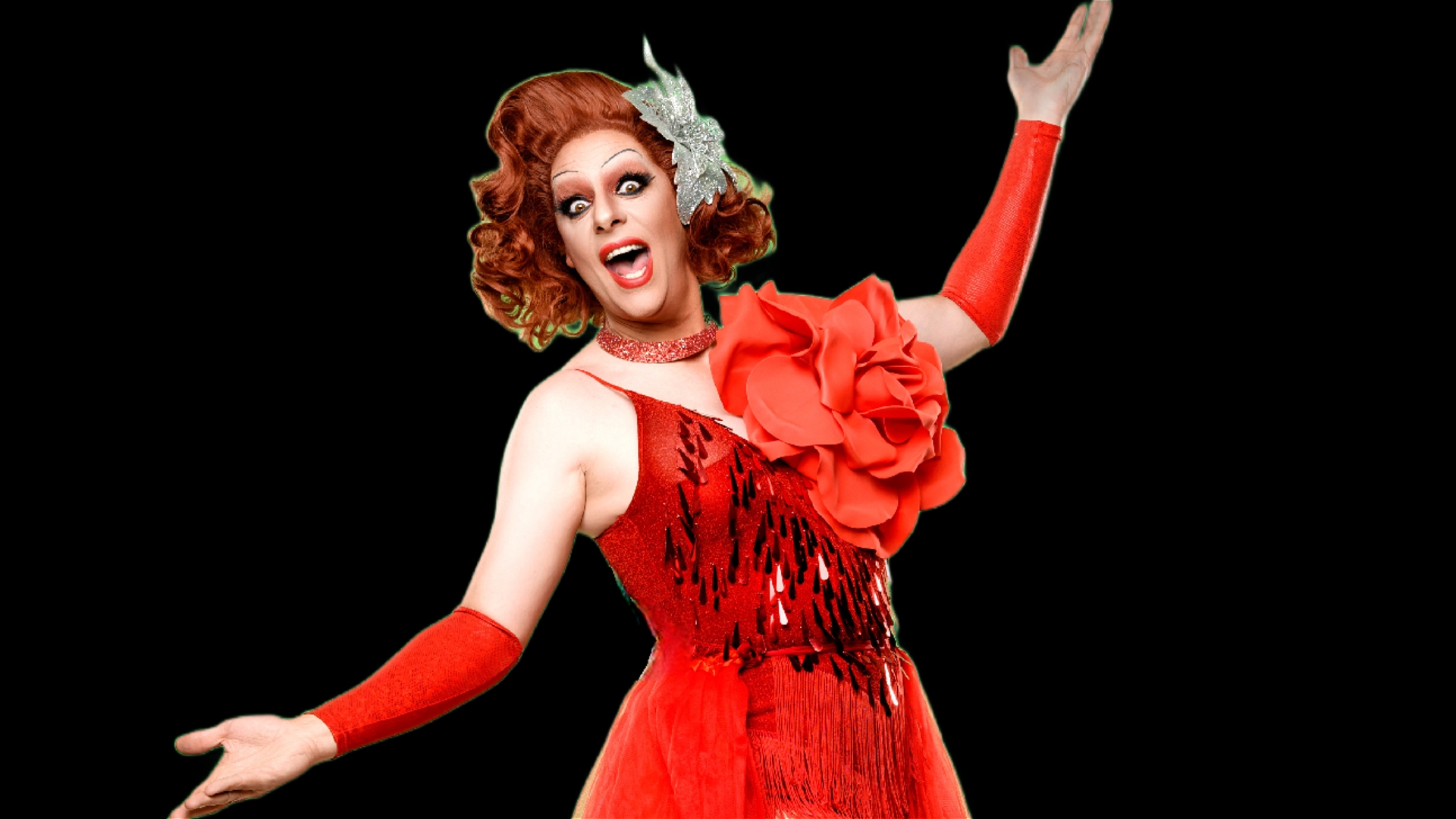 A person in drag wearing a bright red dress with a large flower decoration, red gloves, and a sparkly hair accessory poses with arms outstretched and an expressive, joyful facial expression against a black background.
