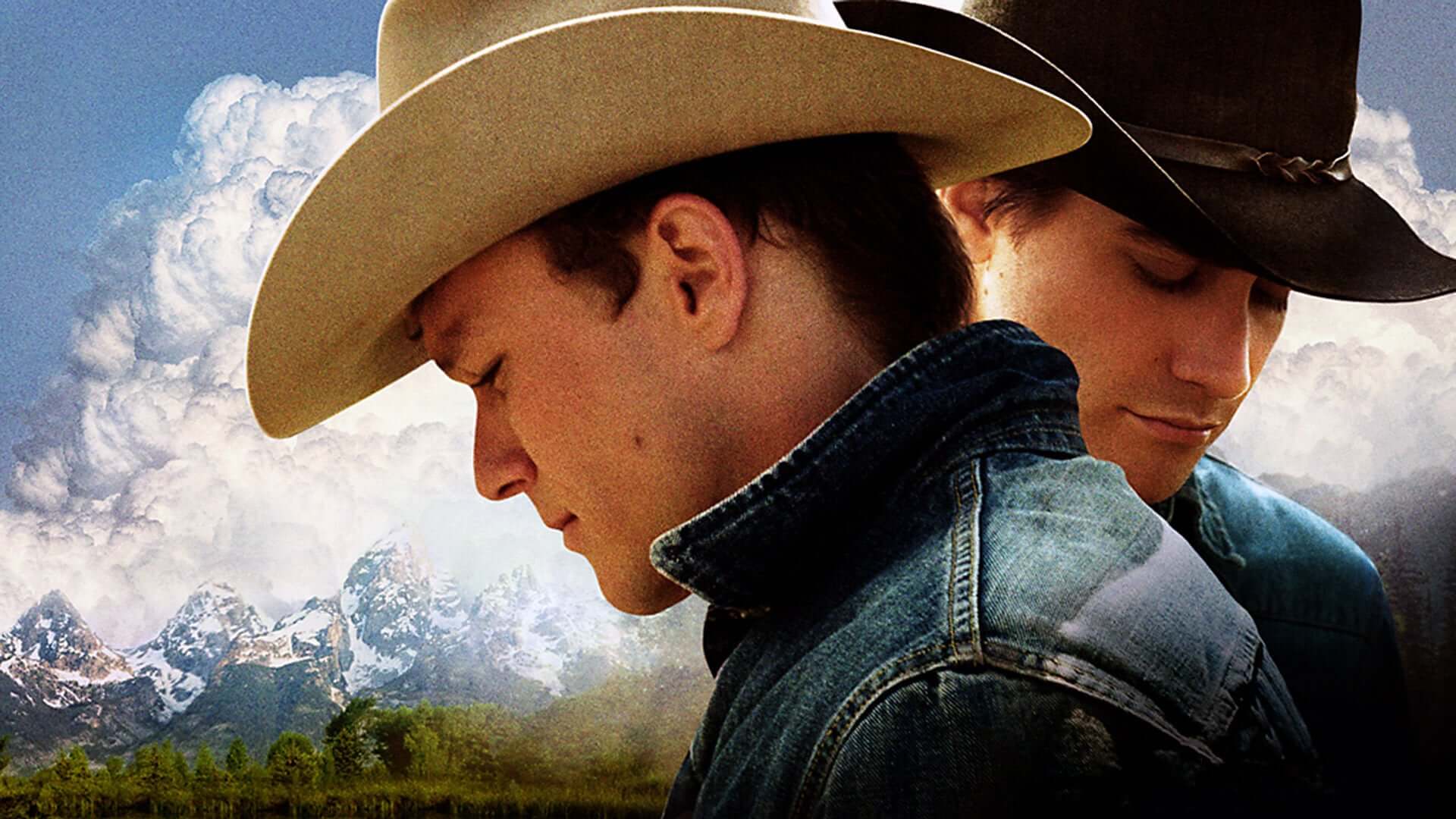 Two men in cowboy hats and denim jackets stand close together outdoors, facing opposite directions with their heads slightly bowed. Snow-capped mountains and a blue sky with clouds appear in the background.