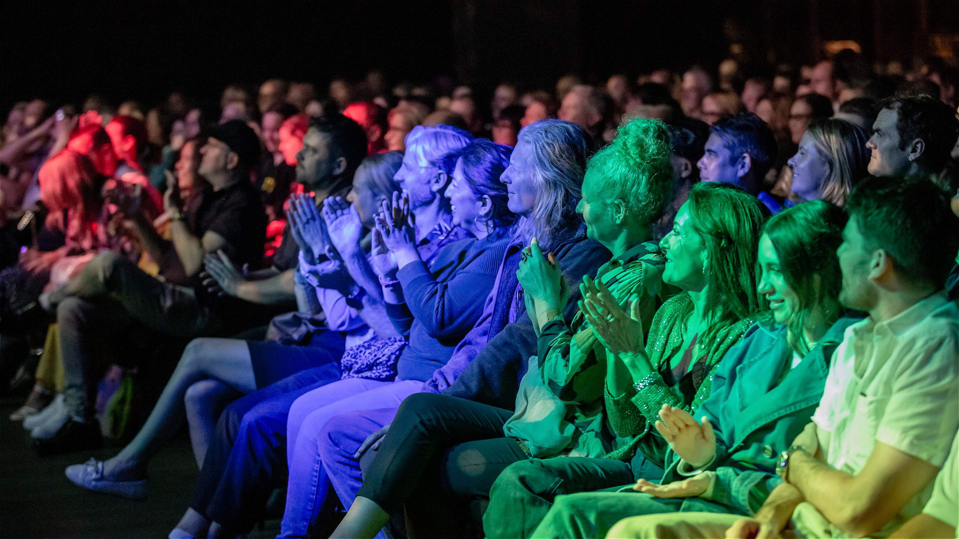 A diverse audience sits closely together in rows, clapping and watching a performance on stage. The crowd is illuminated by colorful stage lights, creating a vibrant atmosphere.