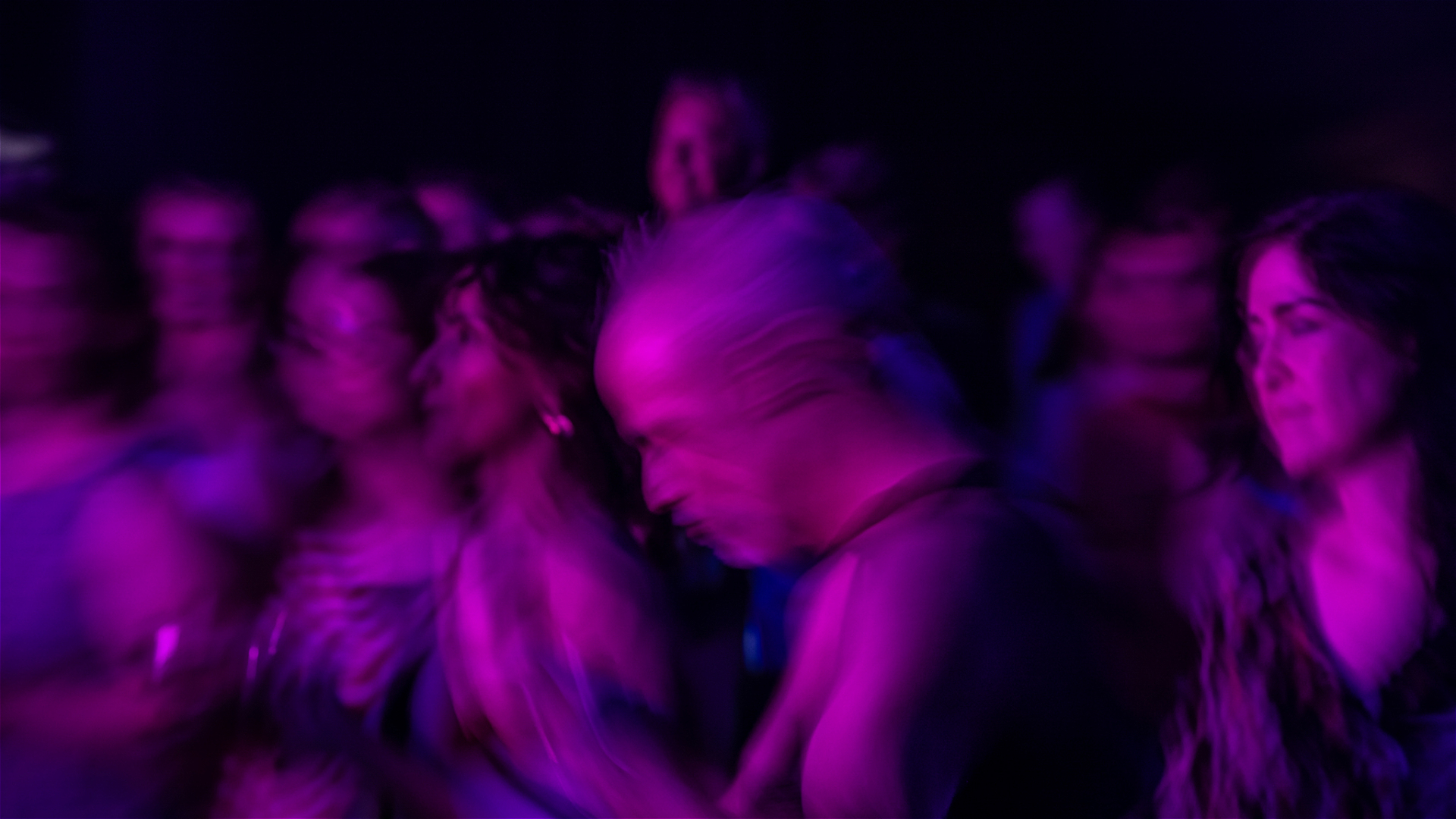 A group of people in a crowd, captured with motion blur and illuminated by purple and pink lights, creating an energetic and dynamic atmosphere.