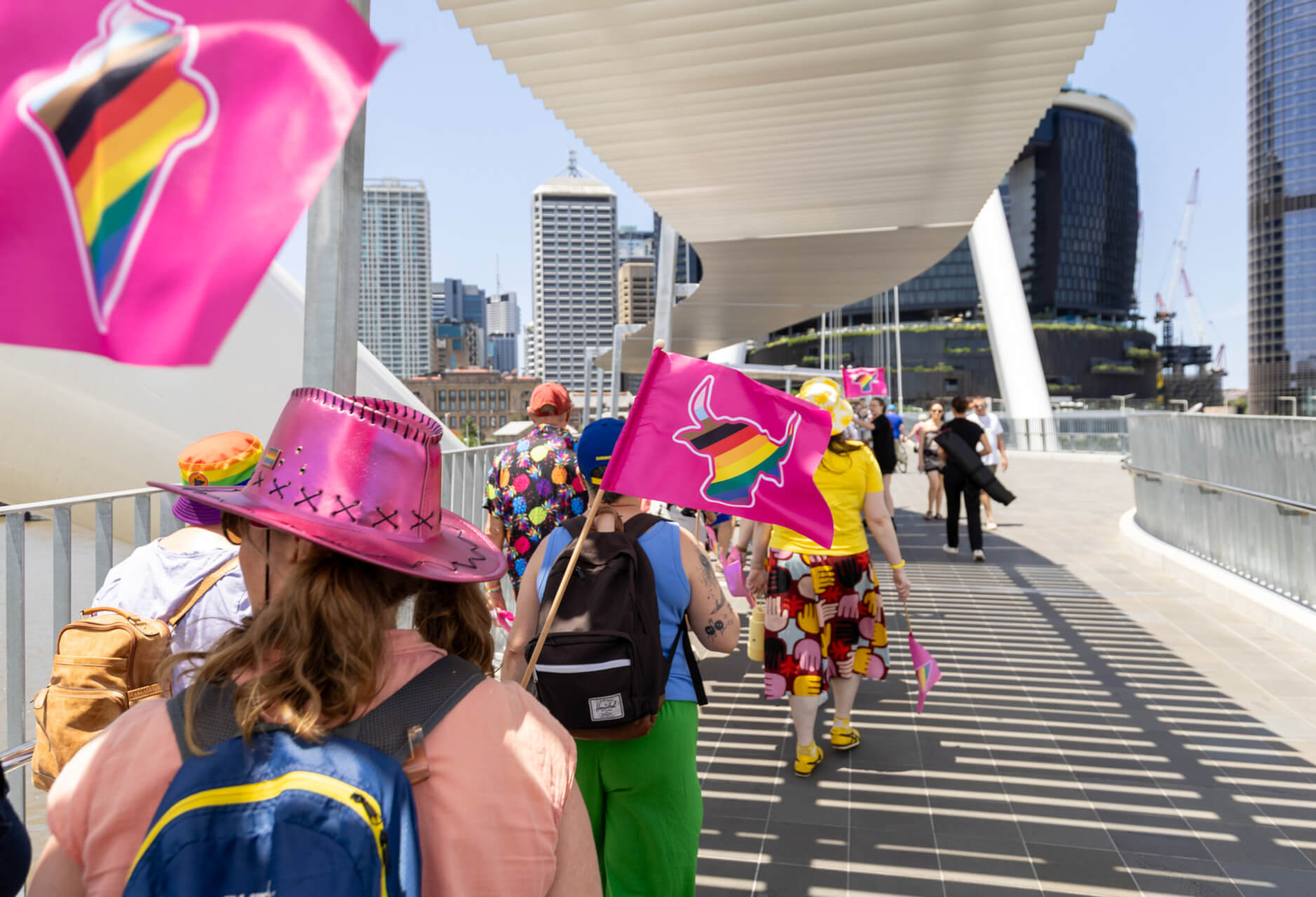 People walk across a city bridge carrying pink flags with a rainbow bullhorn symbol. Some wear colorful clothing and hats, and tall buildings are visible in the background under a sunny sky.