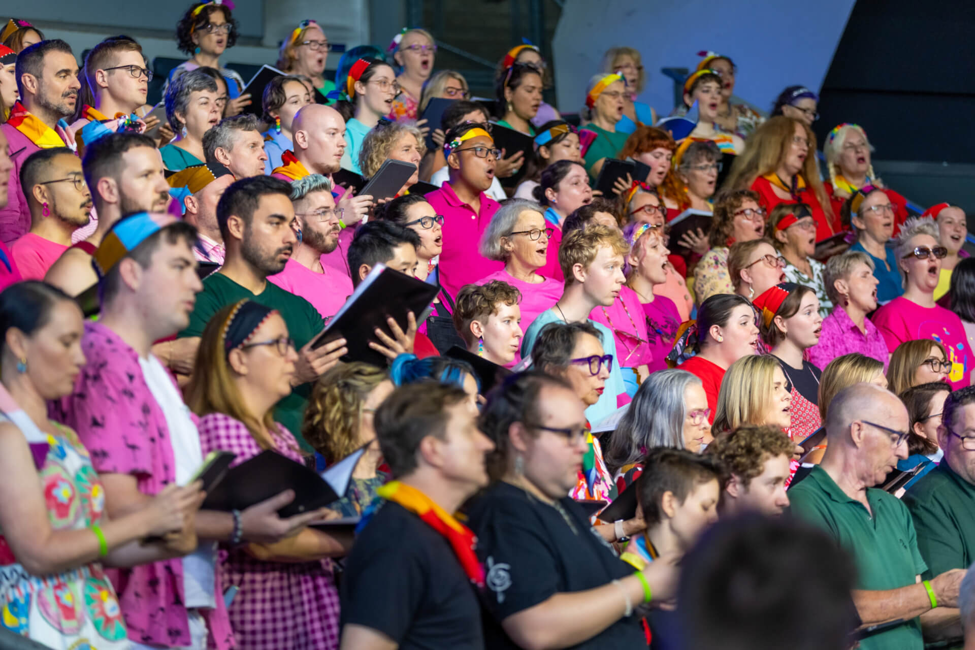 A large, diverse choir sings together, wearing colorful clothing and accessories. Many hold black folders with sheet music, and the group appears joyful and engaged during their performance on stage.