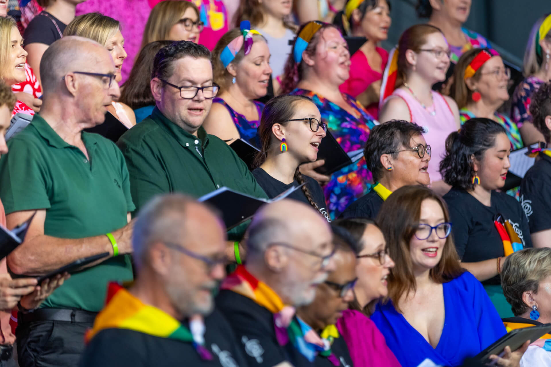 A diverse group of people sing together in a choir, some wearing colorful clothing and rainbow accessories, holding music sheets. The atmosphere is joyful and inclusive.