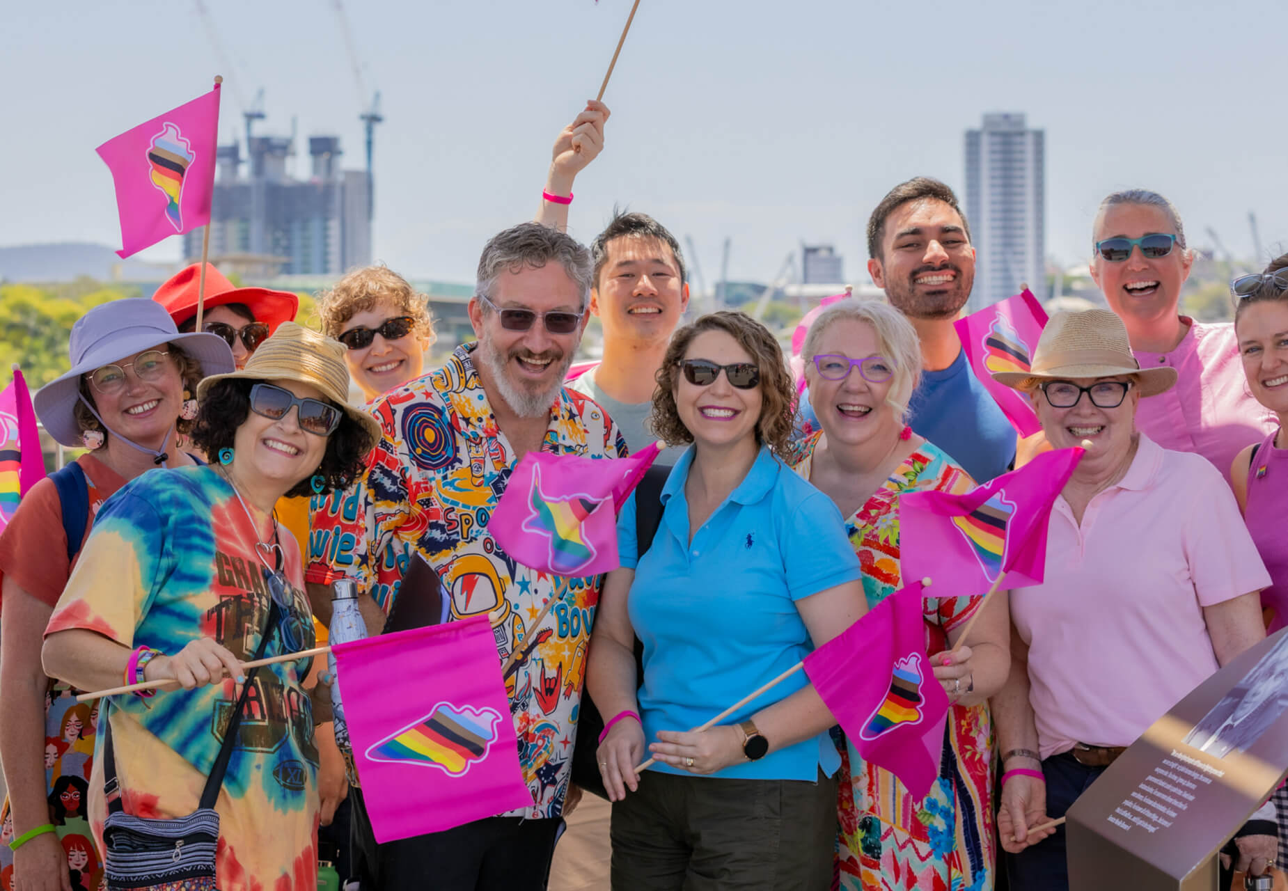 A group of smiling people wave colorful pride flags outdoors on a sunny day, dressed in bright, festive clothing, with city buildings and cranes visible in the background.