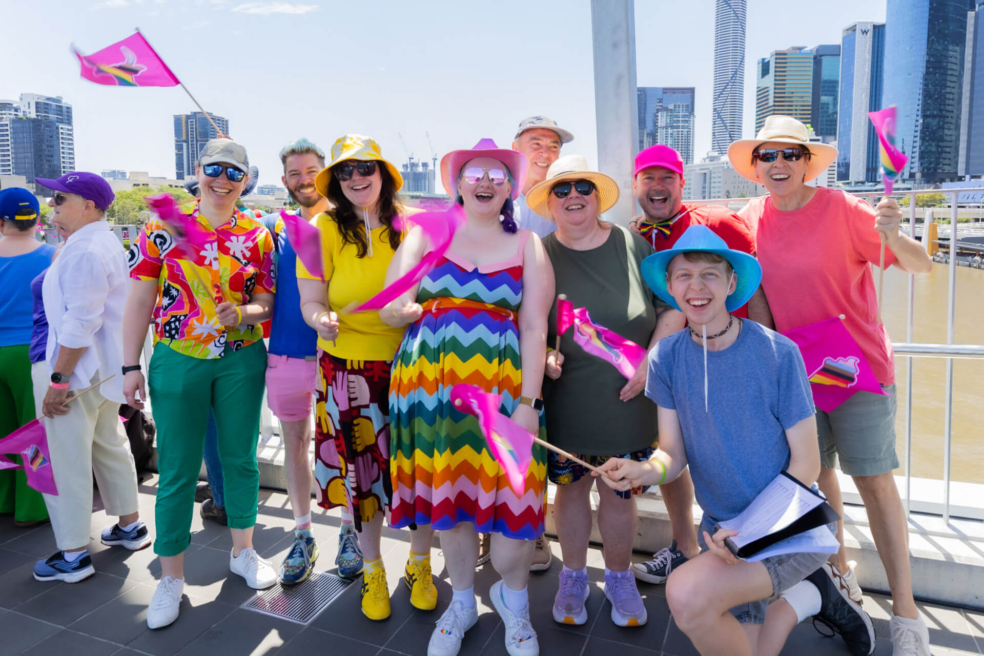 A group of people dressed in colorful, vibrant clothing and rainbow patterns smile and wave small pink flags while standing on a riverside promenade with city buildings in the background on a sunny day.