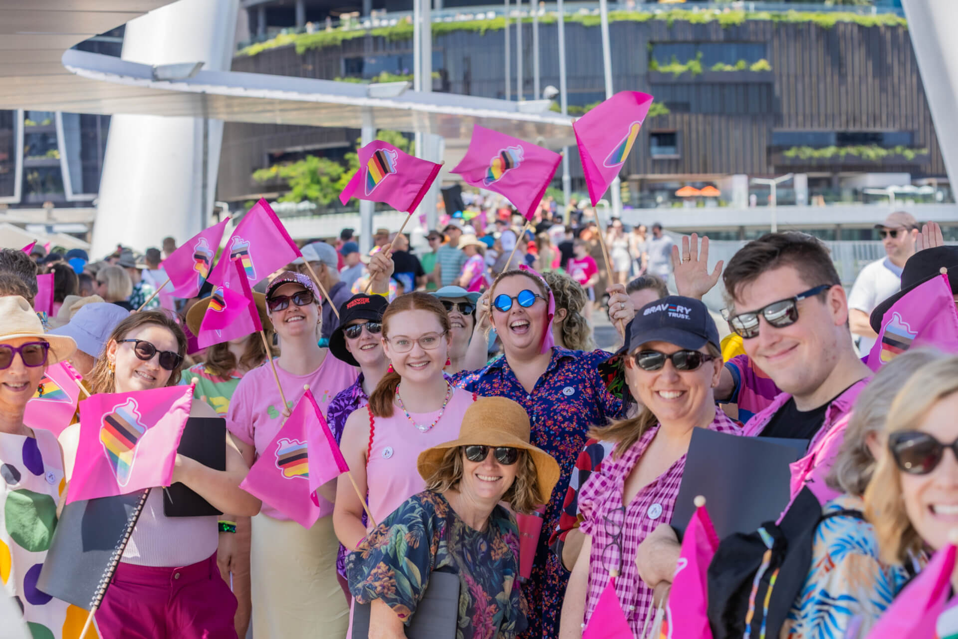 A cheerful group of people wave and smile, holding bright pink flags with cake illustrations, outdoors on a sunny day. Some wear sunglasses and hats; a modern building and bridge are visible in the background.