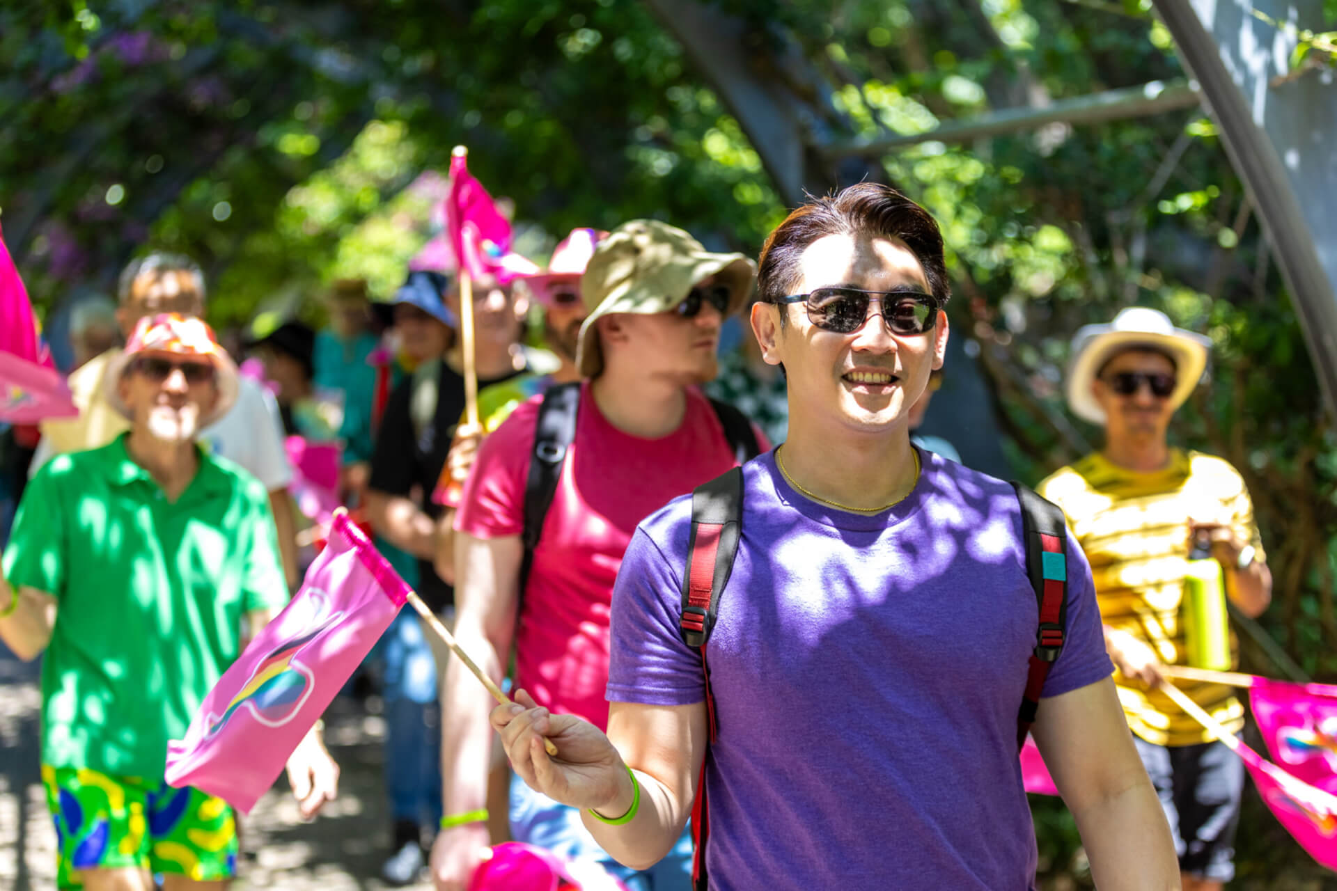 A group of people walk outdoors under leafy trees, smiling and carrying small pink flags. They wear colorful clothes, sunglasses, and hats, enjoying a sunny day together.
