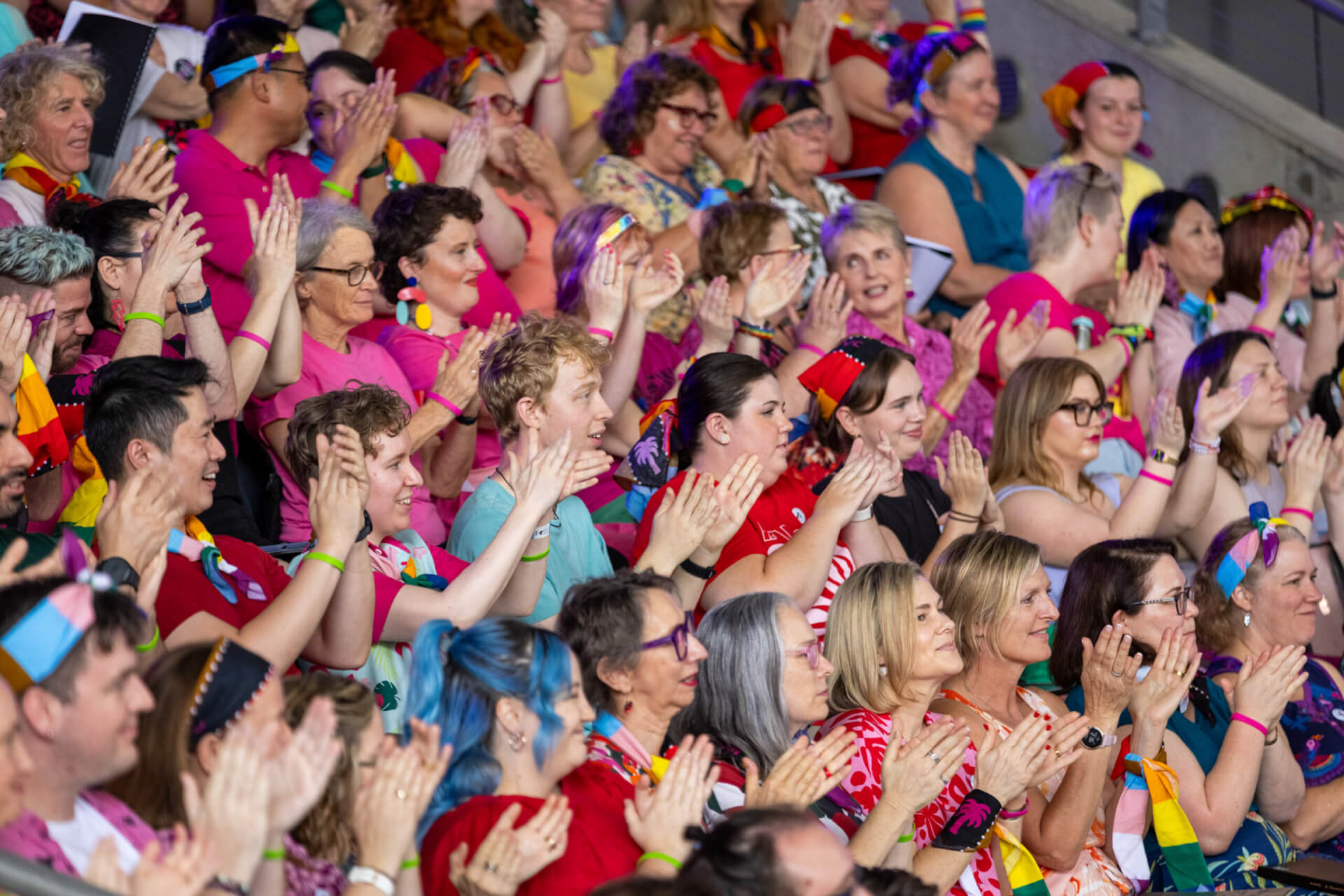 A large, diverse group of people cheer and clap enthusiastically in stadium seating. Many are wearing colorful clothing, rainbow accessories, and headbands, creating a festive and inclusive atmosphere.