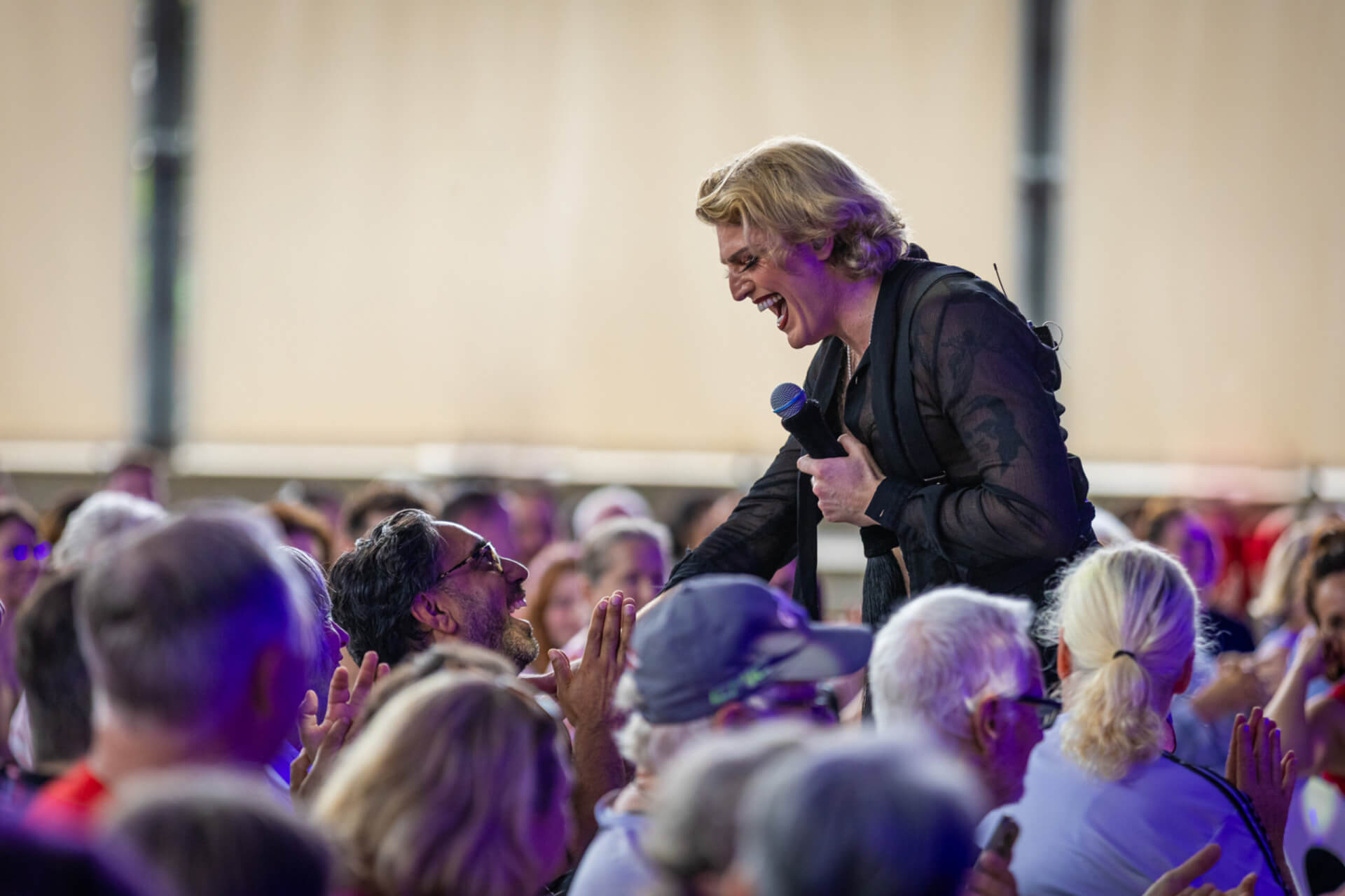 A performer with blond hair and a microphone leans over and smiles at an excited audience member in a crowd, engaging with the lively audience at an indoor event.