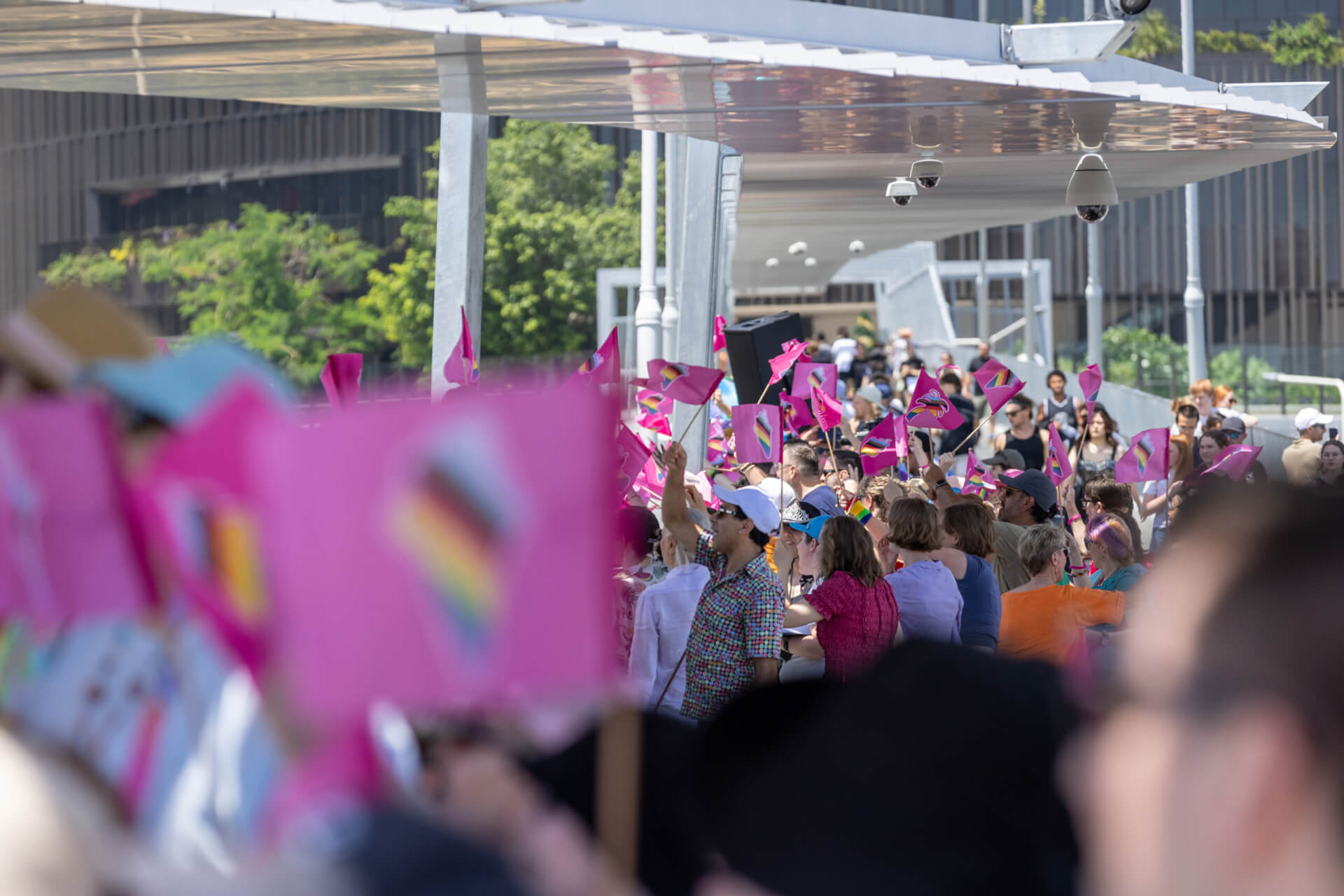 A crowd of people at an outdoor event wave pink flags featuring the Progress Pride flag design. The scene is lively, with attendees standing closely together under a modern, partially covered structure.