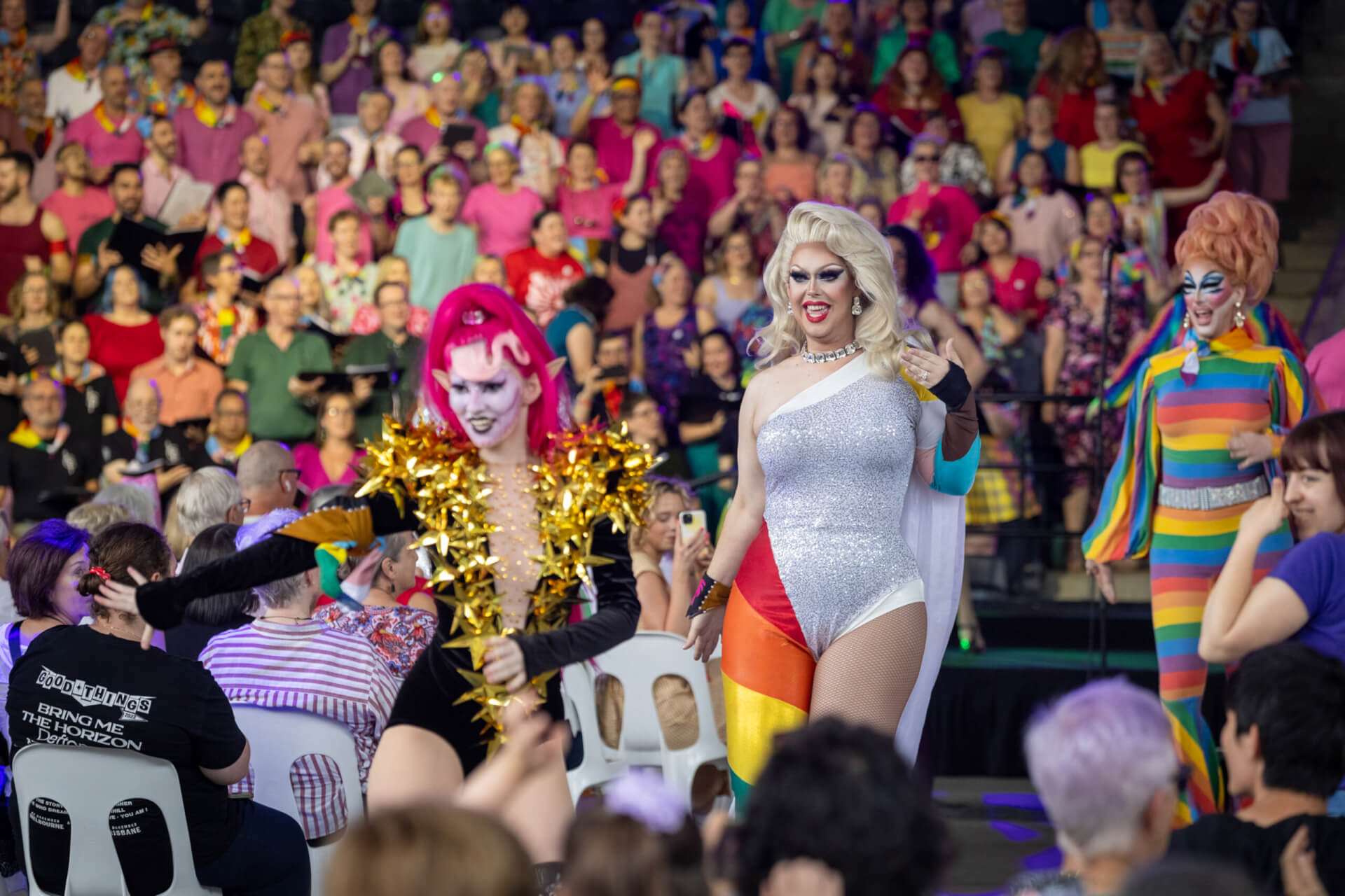 Three drag performers in colorful outfits walk confidently through a crowd. Spectators sit and stand, watching and cheering, with a large group of people in vibrant clothes filling the background.