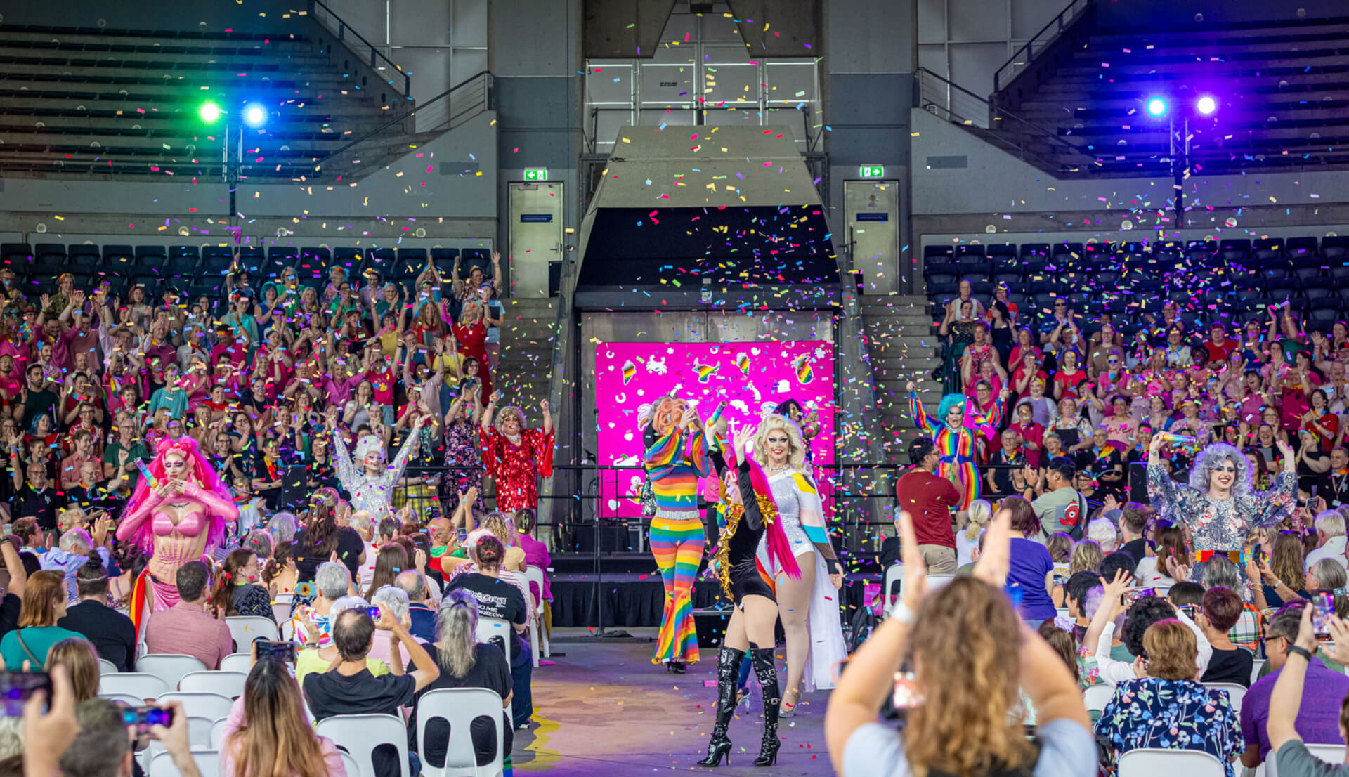 Drag performers in colorful outfits walk down a stage runway as confetti falls, with a cheering audience in a large indoor arena. Bright lights illuminate the scene, creating a festive, celebratory atmosphere.