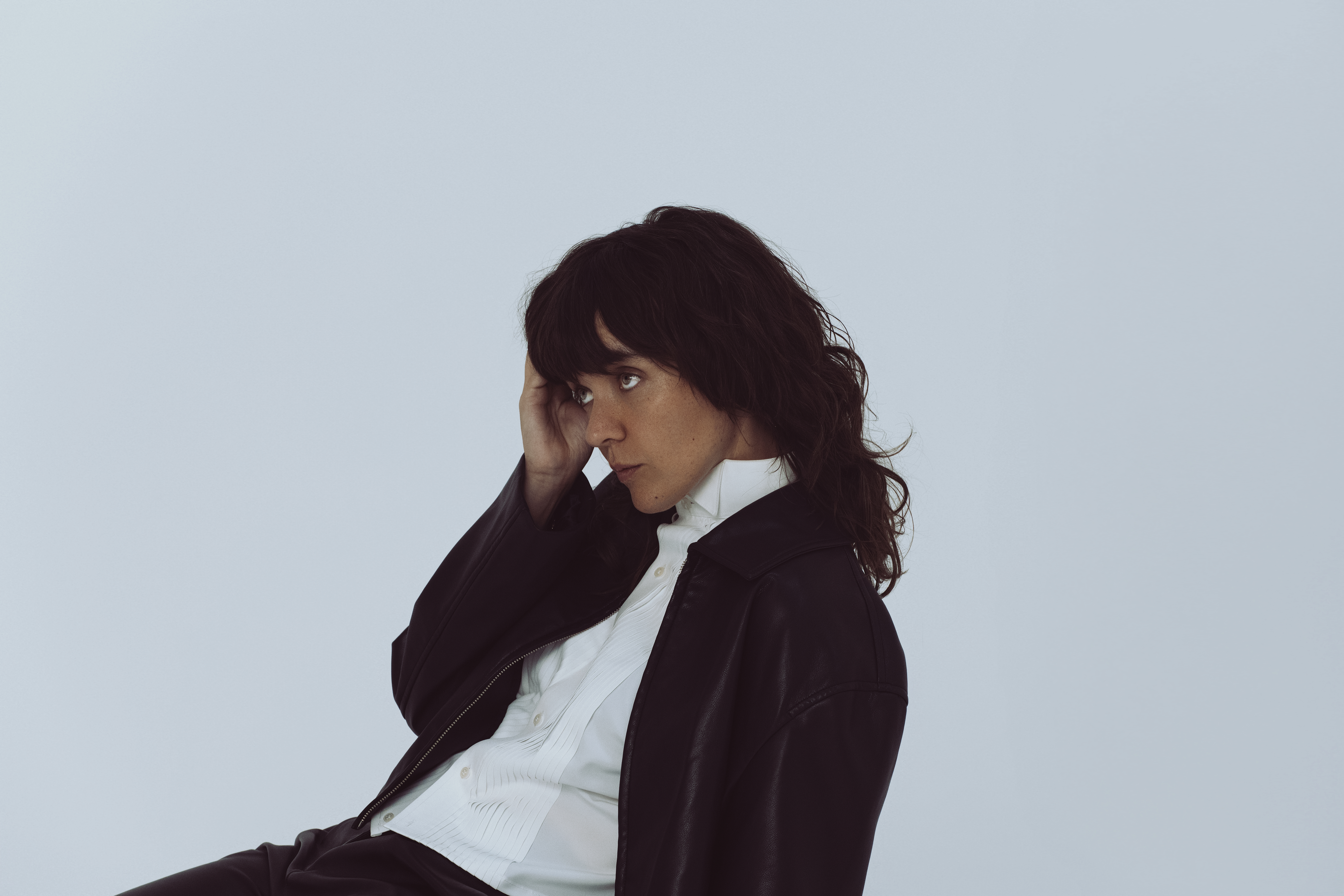 A person with wavy dark hair wears a white shirt and a black jacket, sitting slightly turned with one hand touching their head, against a plain light background.