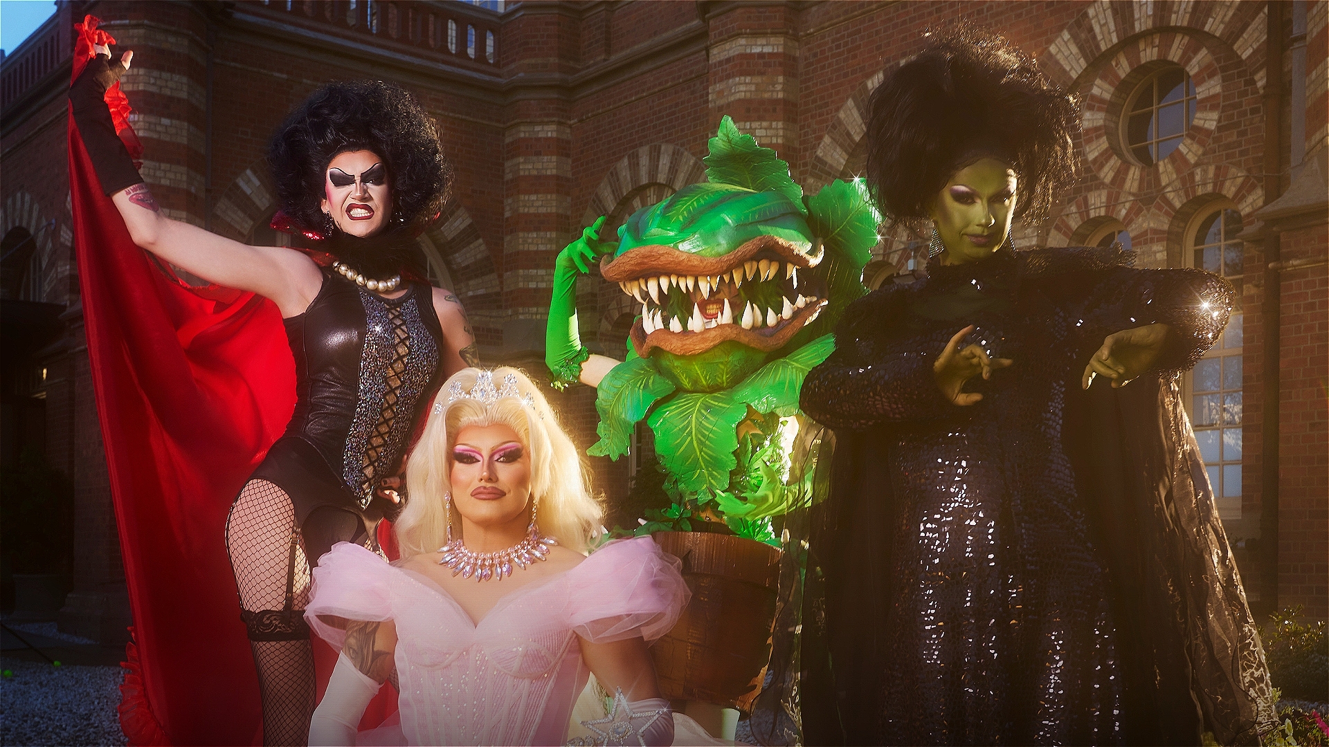 Three drag performers in elaborate costumes and makeup pose with a person in an Audrey II plant costume, standing in front of a brick building with arched windows and dramatic lighting.