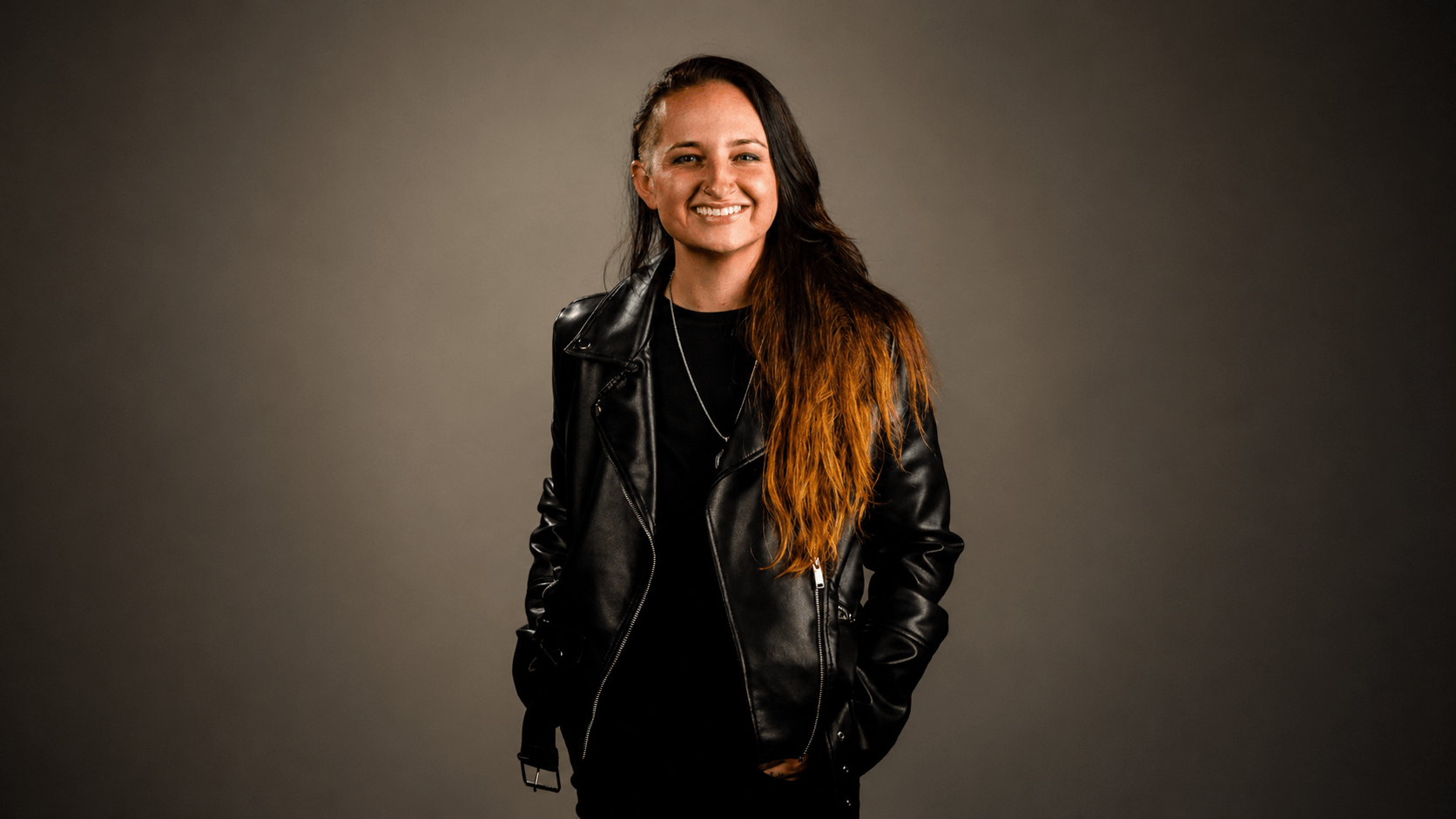 A person with long, partially dyed hair smiles while wearing a black leather jacket and black shirt, standing against a plain, dark background.