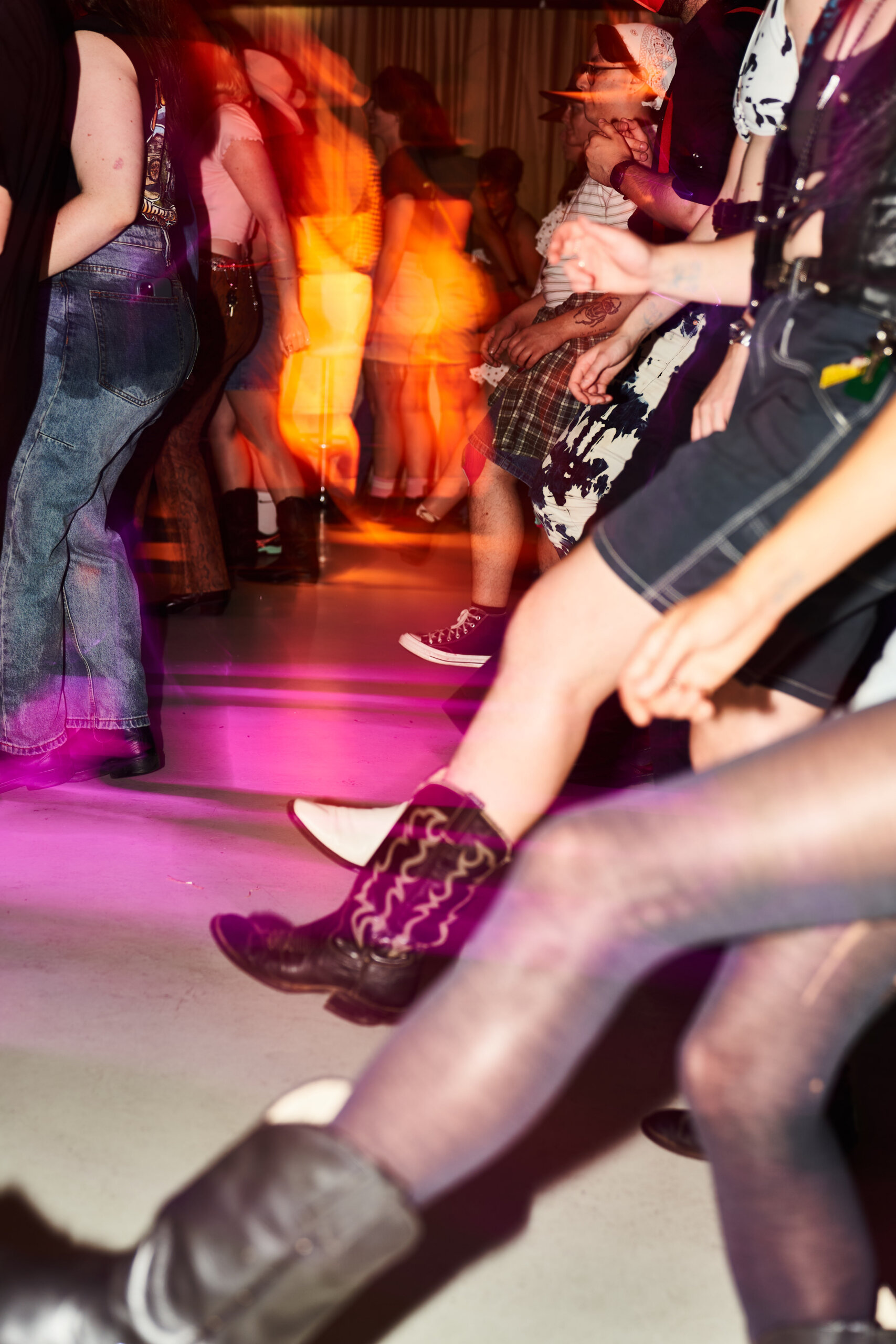 People wearing casual and western-style clothing, including cowboy boots, dance energetically indoors. Motion blur and purple lighting emphasize movement and excitement. Only legs and lower bodies are visible.