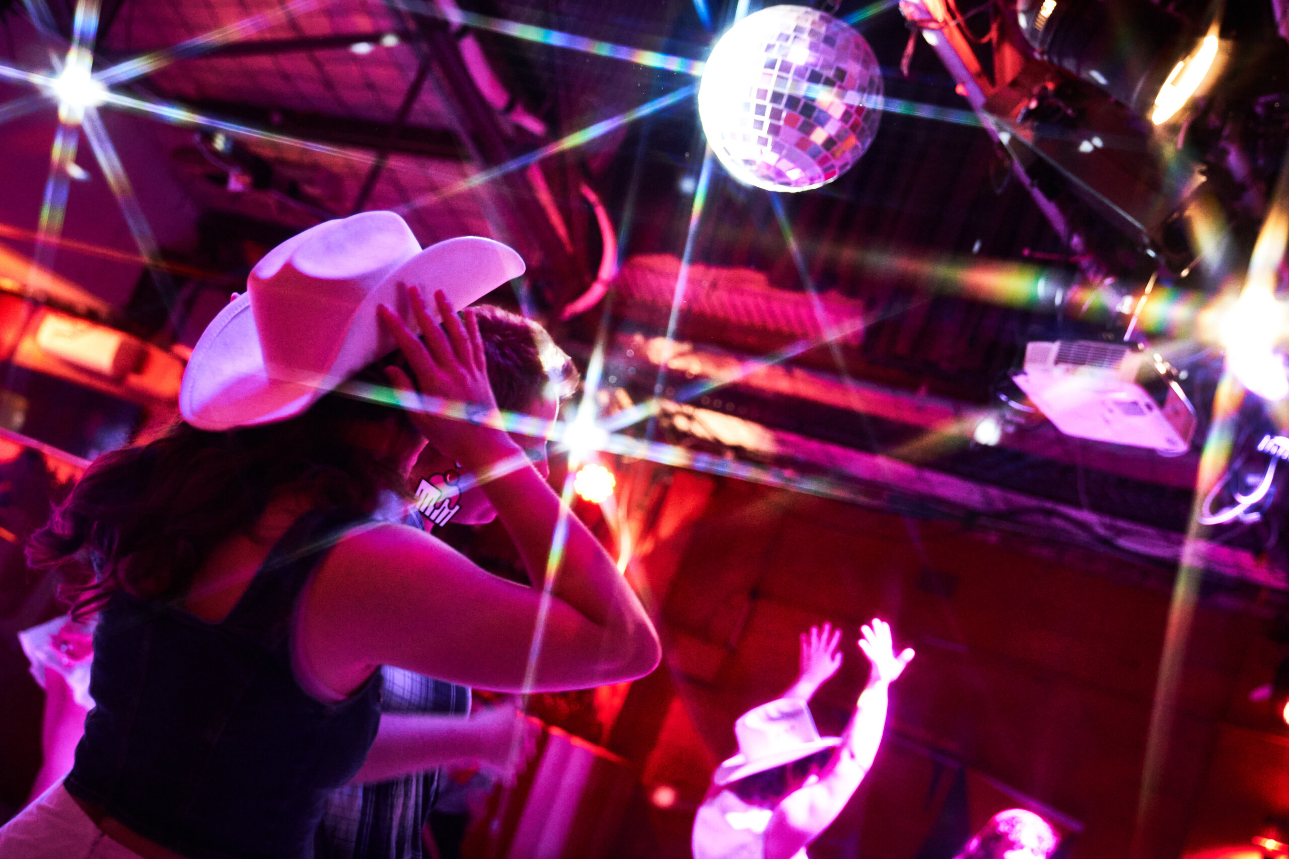People wearing cowboy hats dance under a sparkling disco ball, colorful lights creating starburst effects across the lively, energetic scene.