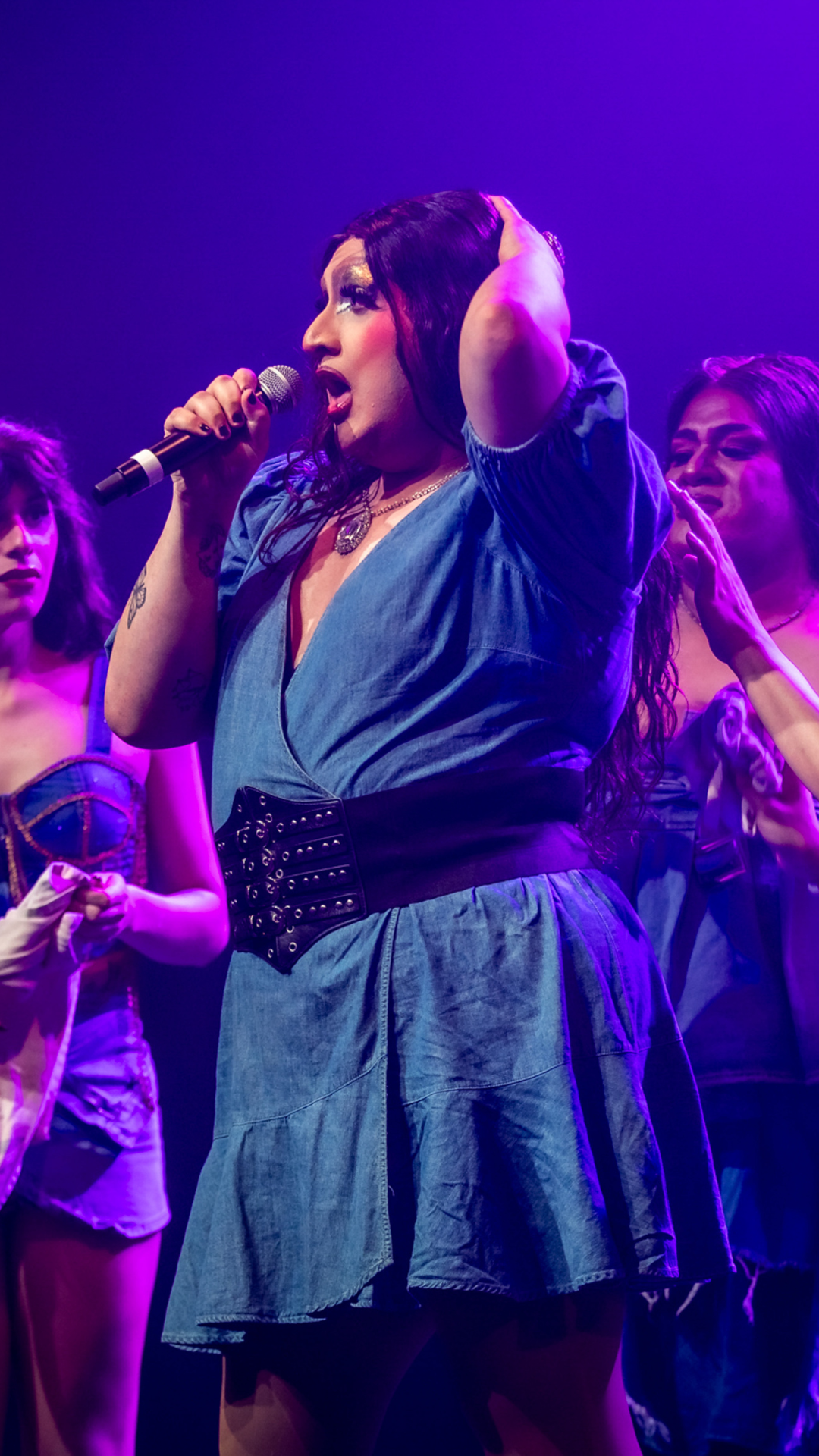 A drag performer in a blue dress sings into a microphone onstage, surrounded by two others with dramatic makeup, under vibrant purple lighting.