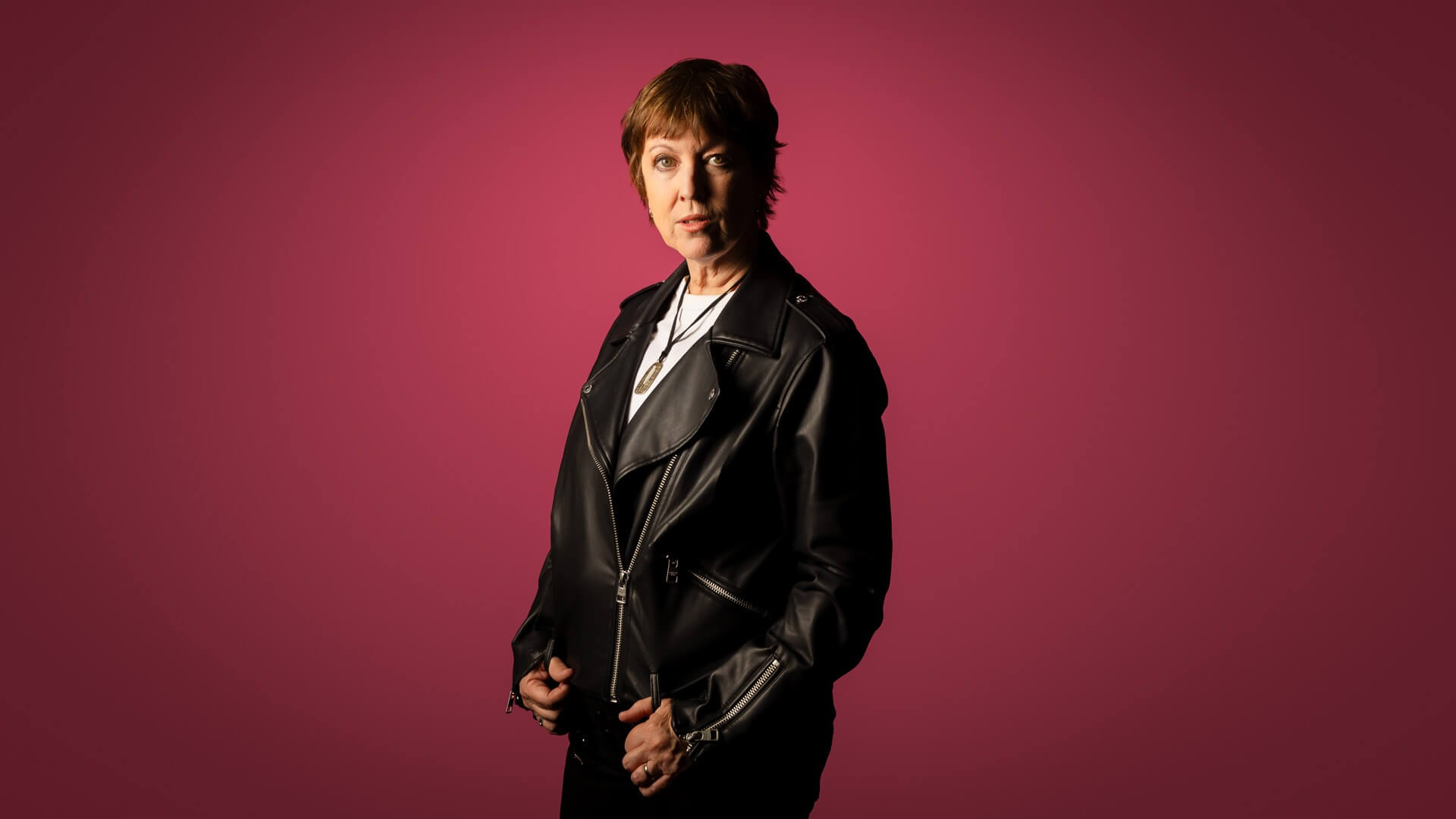 A woman with short brown hair in a black leather jacket stands against a plain pink background, looking confidently at the camera.