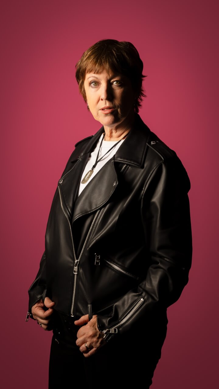 A person with short brown hair, wearing a black leather jacket and white shirt, stands against a solid pink background, looking confidently at the camera.
