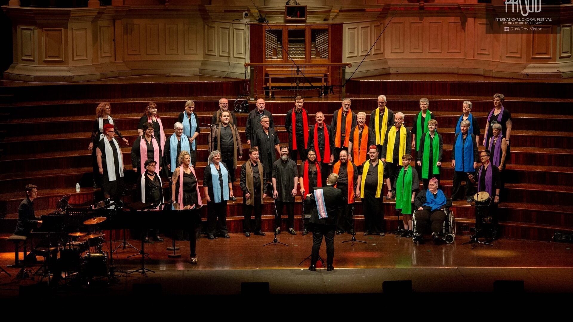 A choir of men and women stands on stage in tiers, each wearing a black outfit with a brightly colored scarf. A conductor faces the group. Musical instruments are visible at the left of the stage. The background is a wooden concert hall.