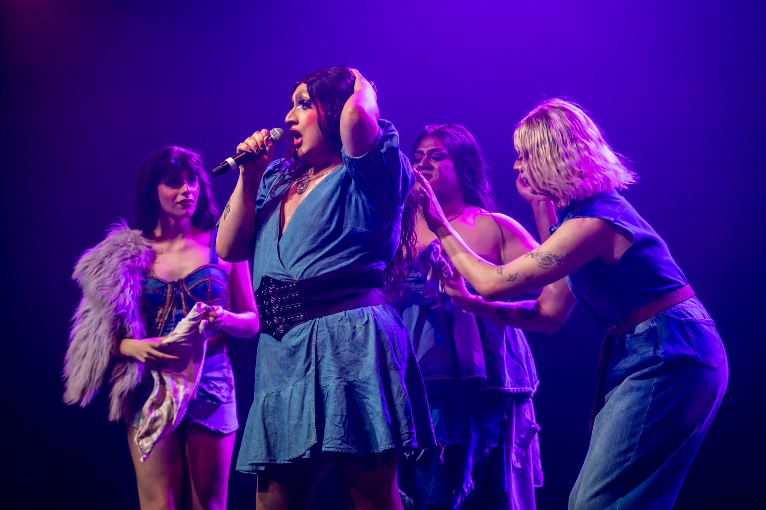 Four performers in blue outfits stand on stage under purple lights. One sings into a microphone while the others interact with her, adjusting her hair and clothing. The atmosphere is lively and theatrical.