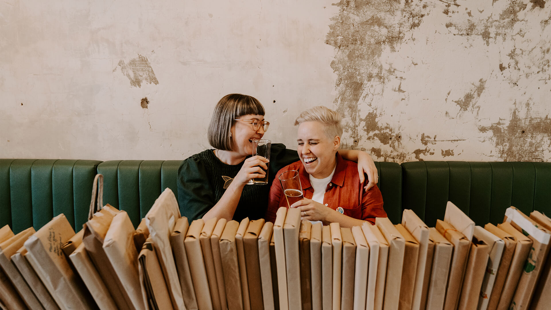 Two people sit together in a green booth, laughing and holding drinks, with wrapped books stacked on the table in front of them against a worn, textured wall.