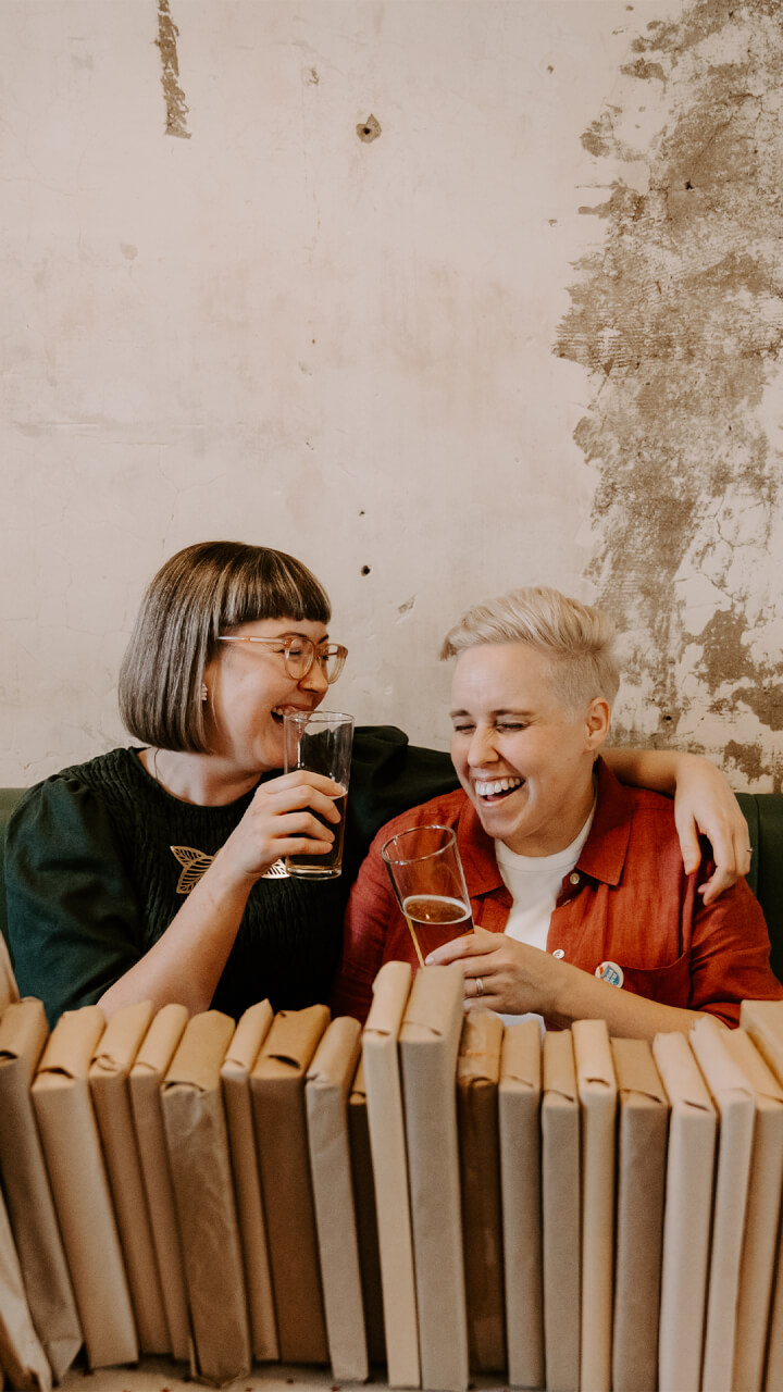 Two people sit close together, laughing and holding drinks, with one arm around each other. They are in front of a rustic wall and a row of books wrapped in brown paper.