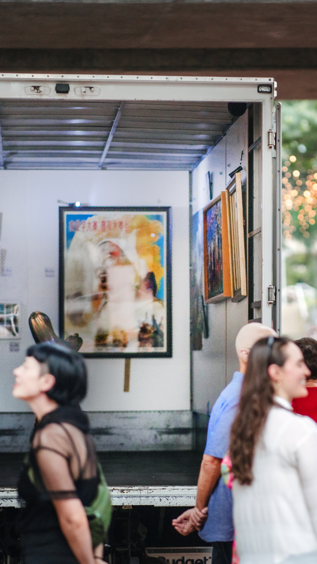 A group of people stand near an open truck displaying various framed artworks, including a large, colorful portrait at the center, with trees and lights in the blurred background.