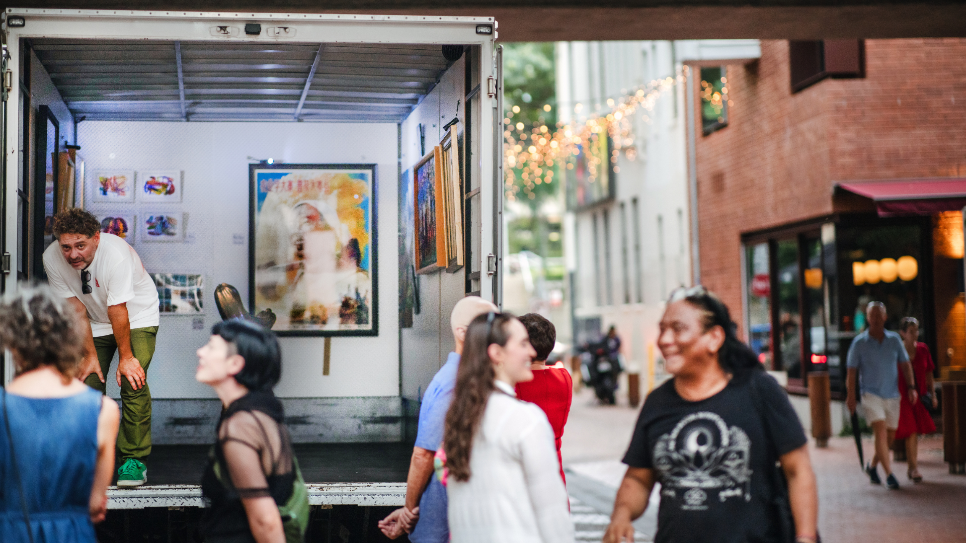 People gather around an open truck displaying artwork inside, with one person standing in the truck. The scene is lively and takes place on a city street in the evening, with lights and buildings in the background.