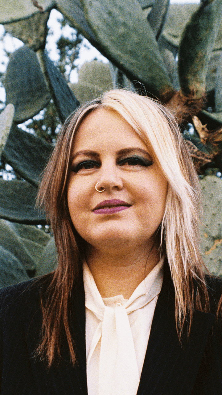 A person with long, two-toned blonde and brown hair, bold makeup, and a nose ring smiles slightly. They wear a white blouse and dark blazer, standing in front of large cactus pads outdoors.