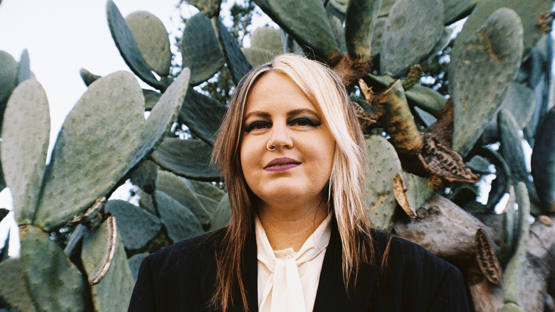 A person with long, straight hair and bold makeup stands in front of large prickly pear cactus pads, wearing a black blazer and light-colored blouse, outdoors on a bright day.