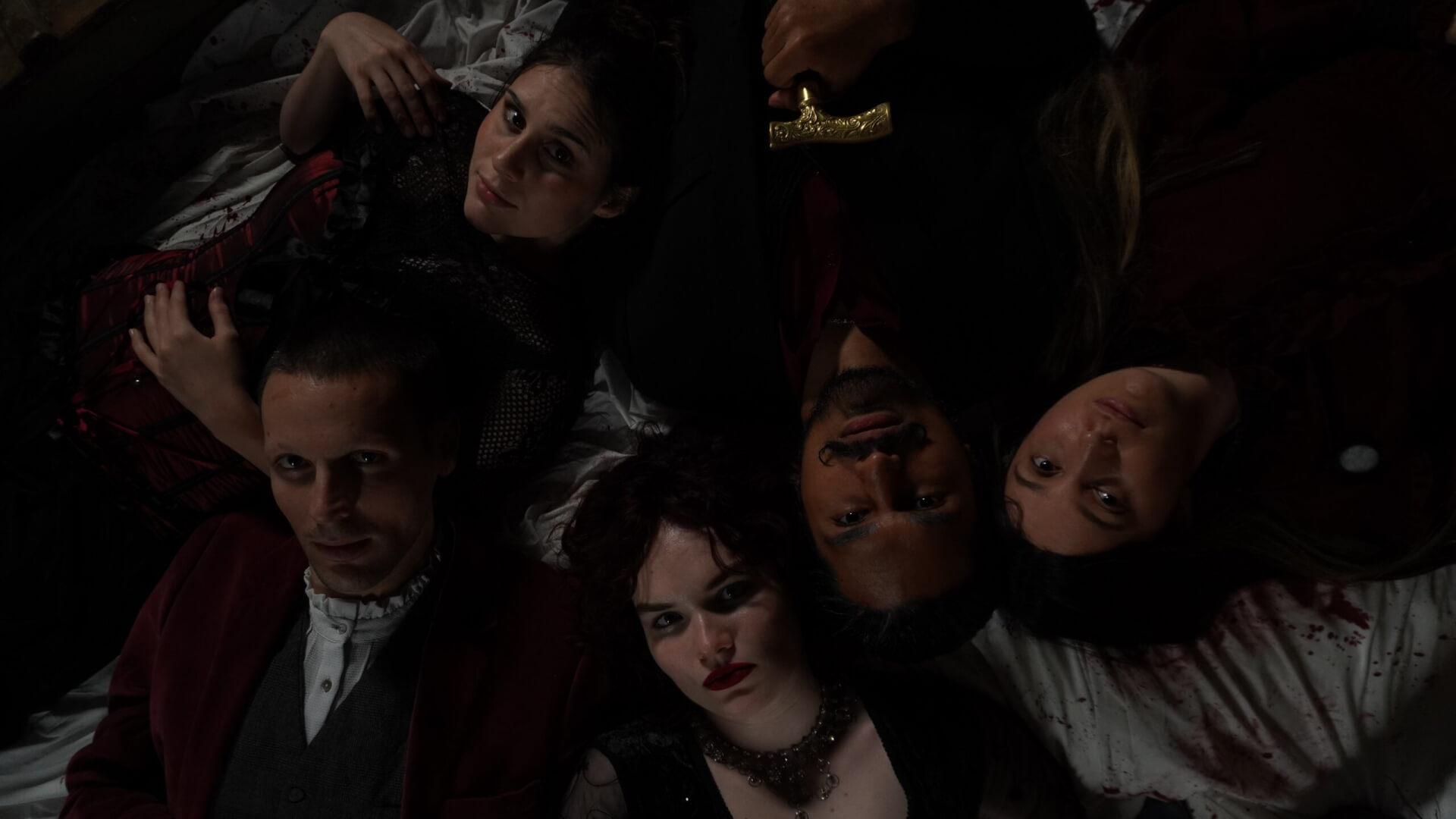 Five people in dark, gothic-style clothing lie on a bed, heads close together, looking up at the camera. The scene is dimly lit, creating a moody and dramatic atmosphere.