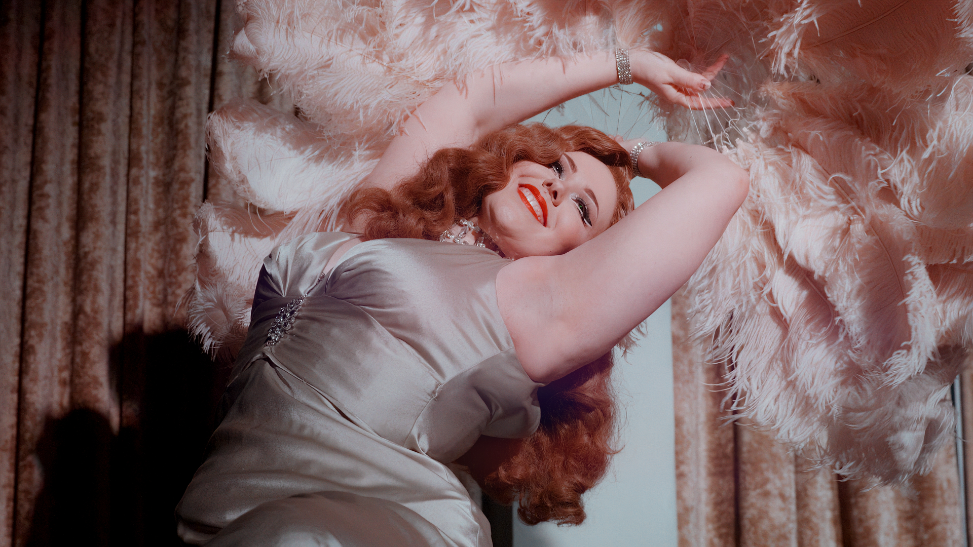 A woman in a satin gown poses dramatically, smiling and holding large pink feather fans above her head, with long red hair and red lipstick, standing in front of beige curtains.